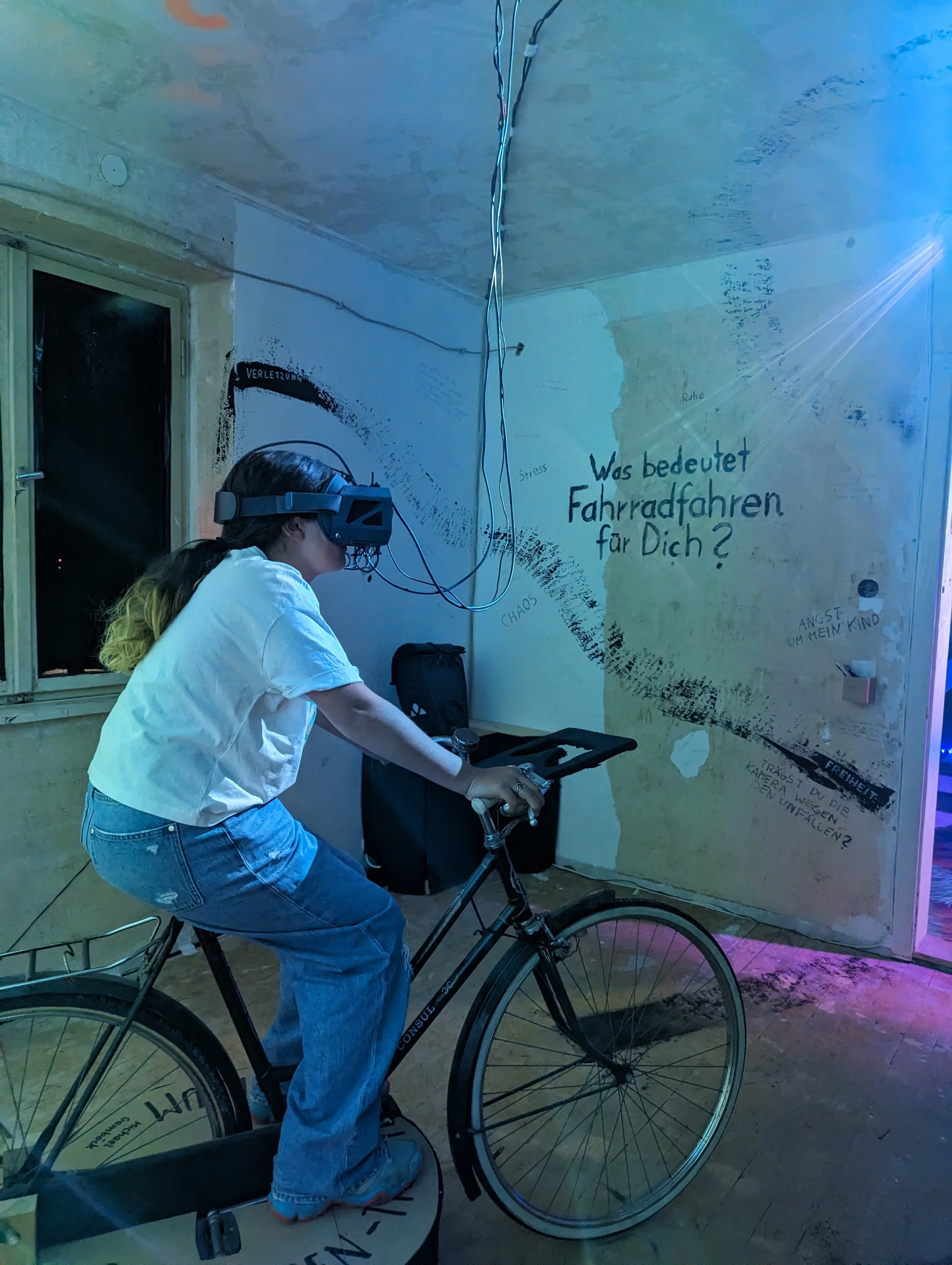 A woman is riding a stationary bicycle equipped with a virtual reality headset, in a room with graffiti and writings on the walls, including a question in German asking about the meaning of riding a bike for you.