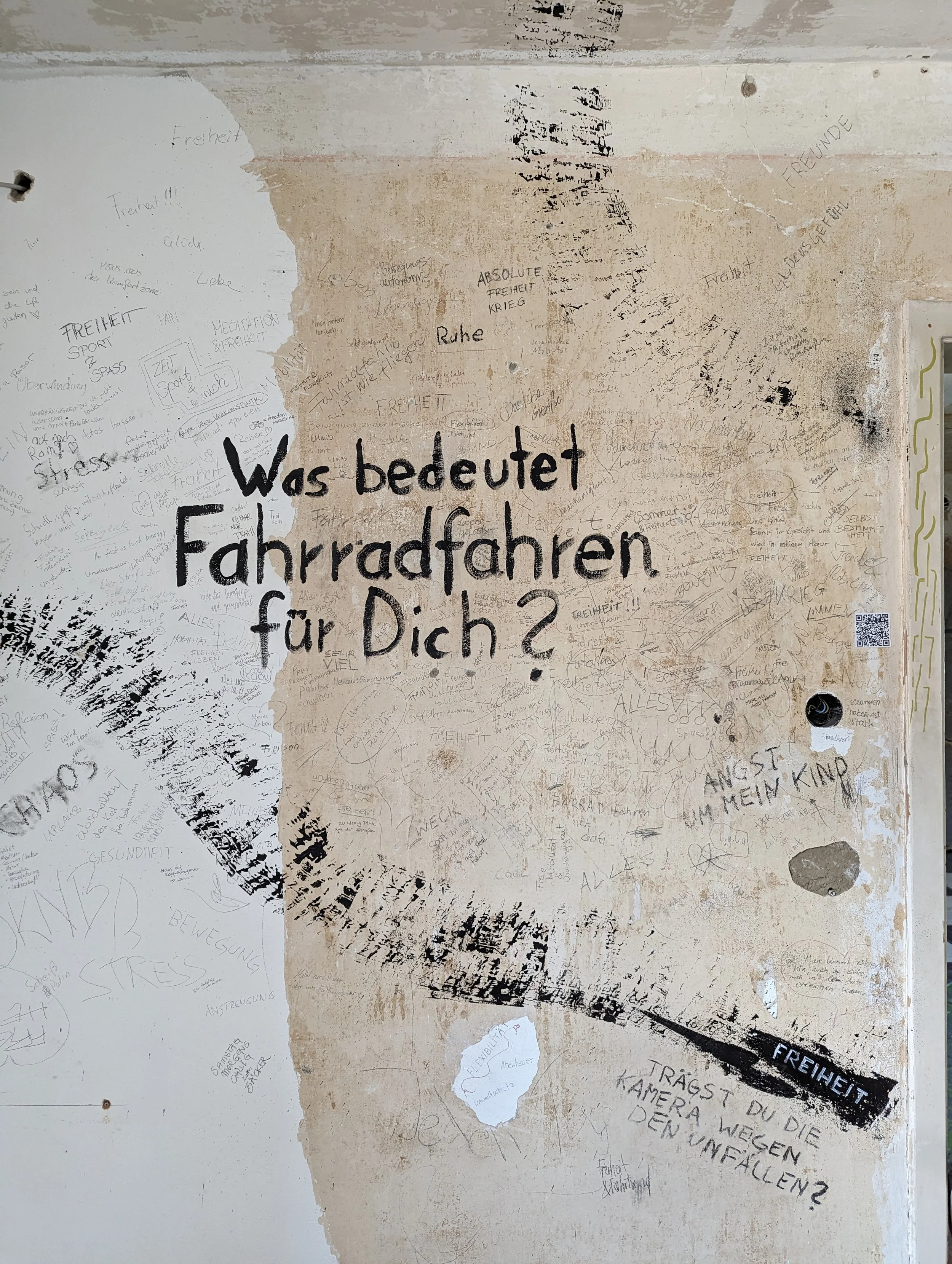 A wall with graffiti and writing, including the German question 'Was bedeutet Fahrradfahren für Dich?' in large black letters, and other handwritten messages and drawings in various colors and styles.