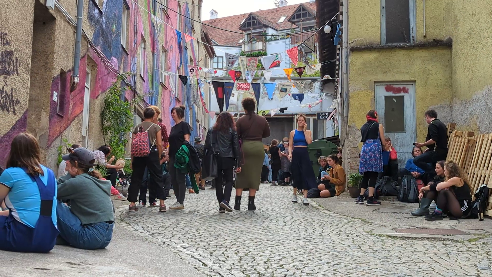 People walking and sitting in a narrow, cobblestone street decorated with colorful bunting, murals, and graffiti art on the walls.