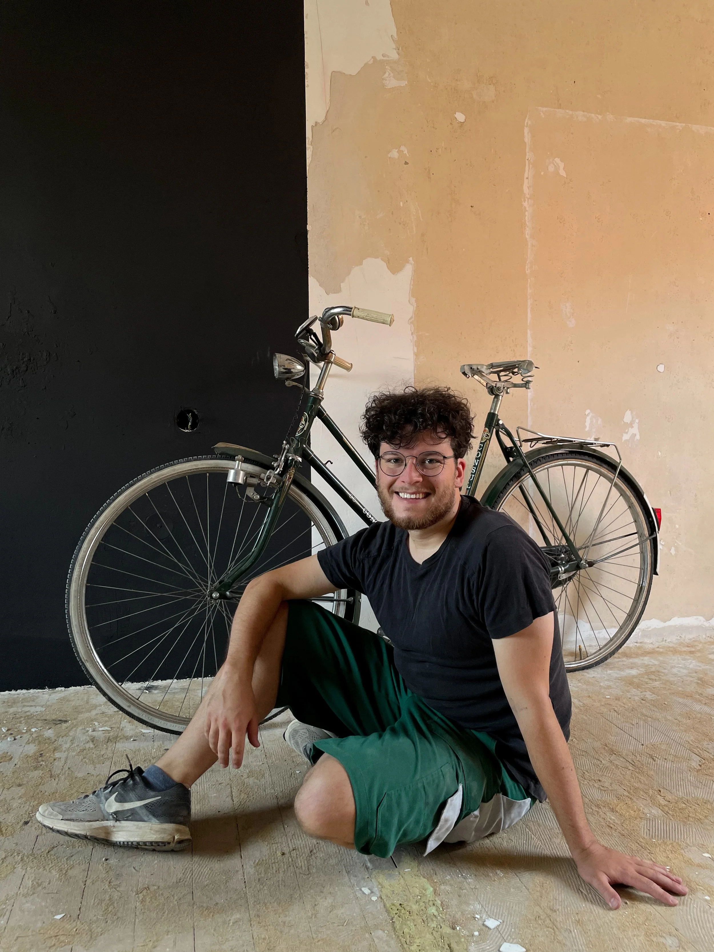 Michael Gamböck with curly hair, glasses, and a beard sitting on the floor in front of a vintage bicycle, smiling at the camera, with a partially painted wall in the background.