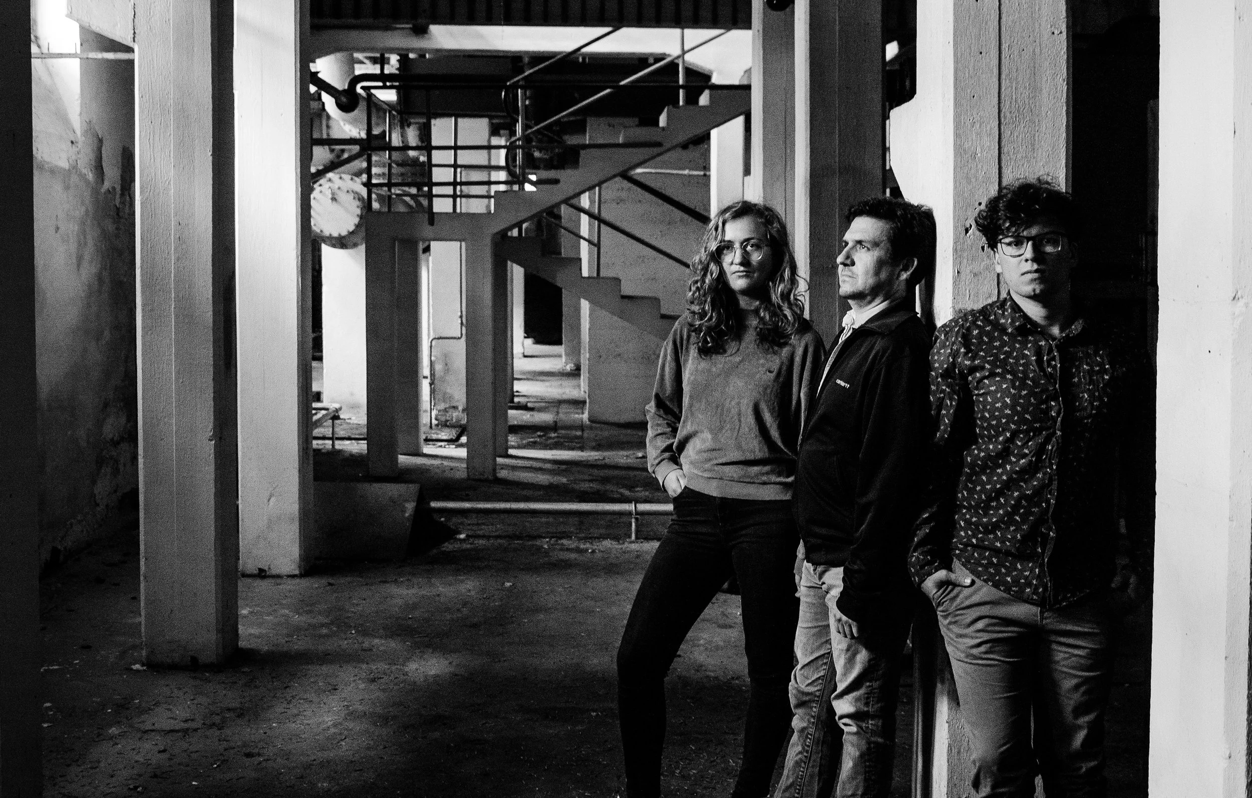 Franziska Hauber, Tom Simonetti and Michael Gamböck stand against a wall in an unfinished industrial space with exposed beams and a staircase in the background, in black and white.