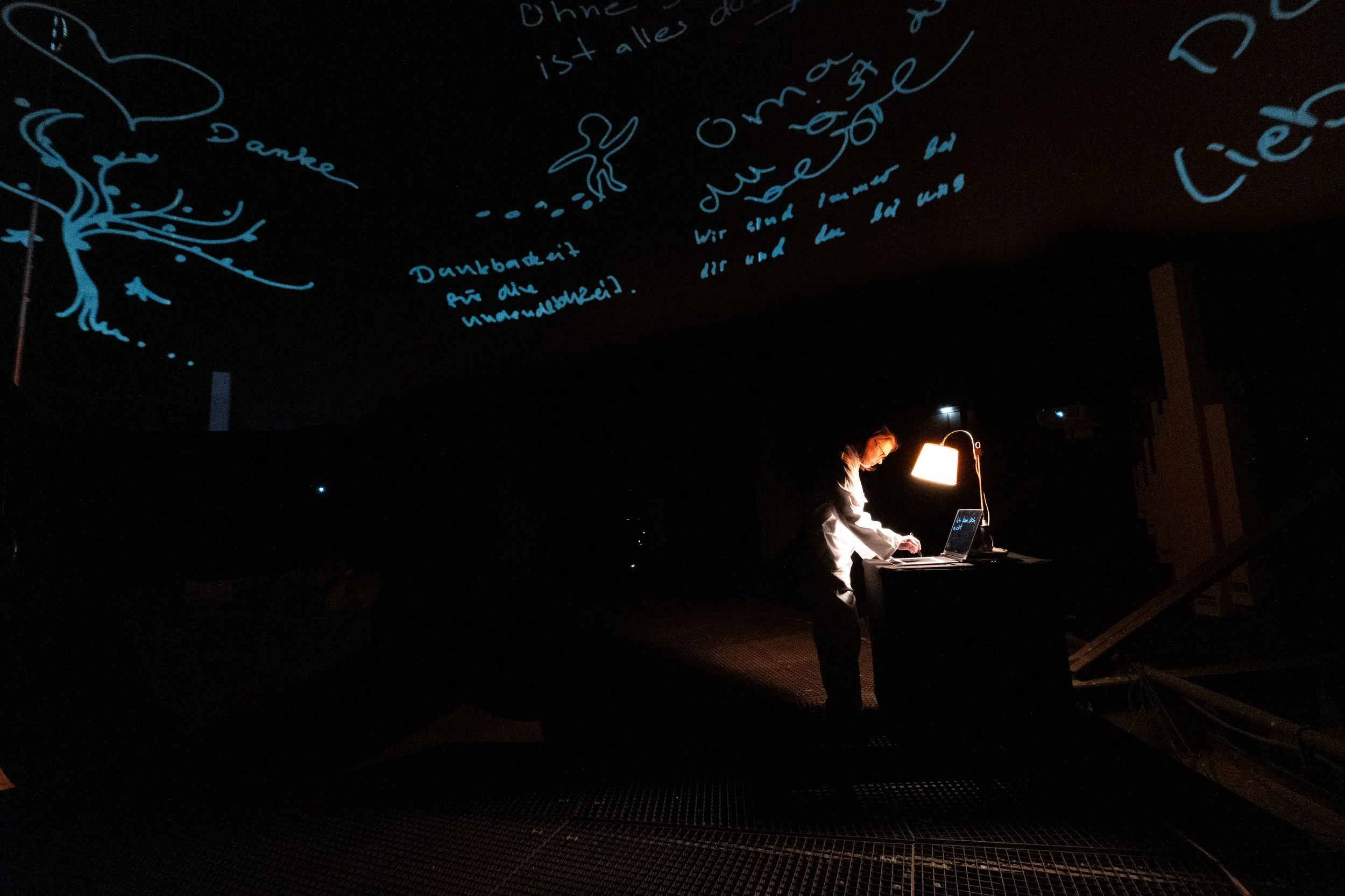 A person working at a desk illuminated by a desk lamp, with dark surroundings and handwritten notes projected or written on the ceiling in blue ink.