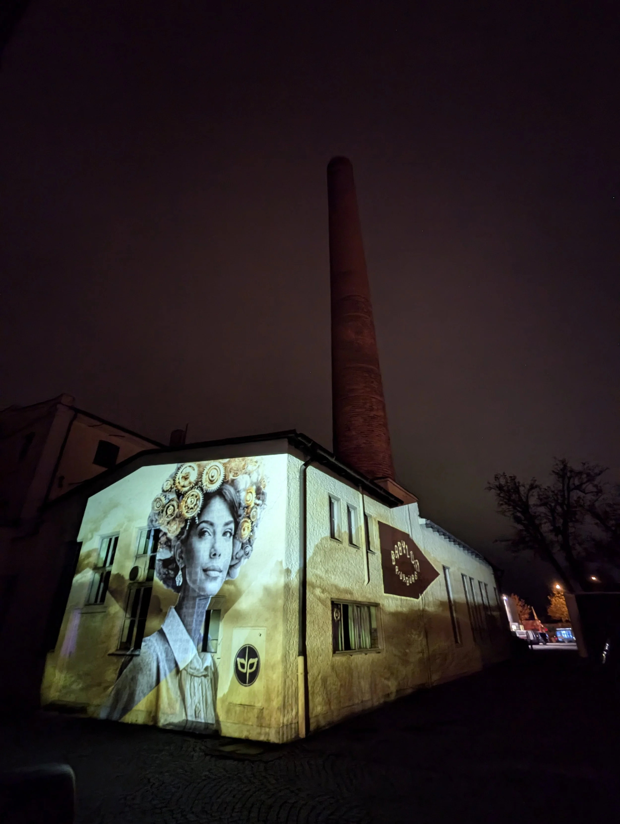 A building at night with a projection of a black and white woman with curly hair decorated with flowers on its corner wall, and a tall smokestack in the background.
