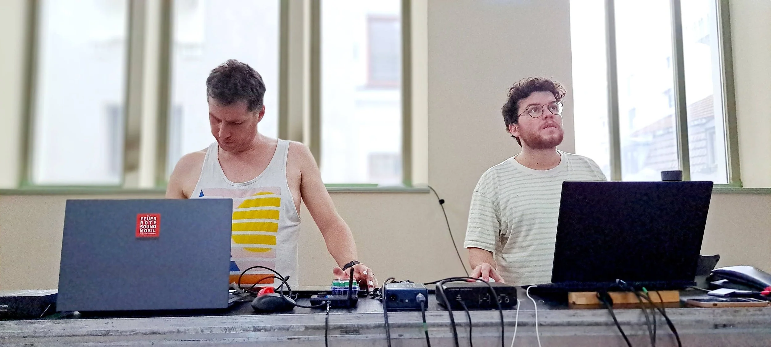 Two men working with electronic music and light equipment at a table in an old bathhouse with large windows.