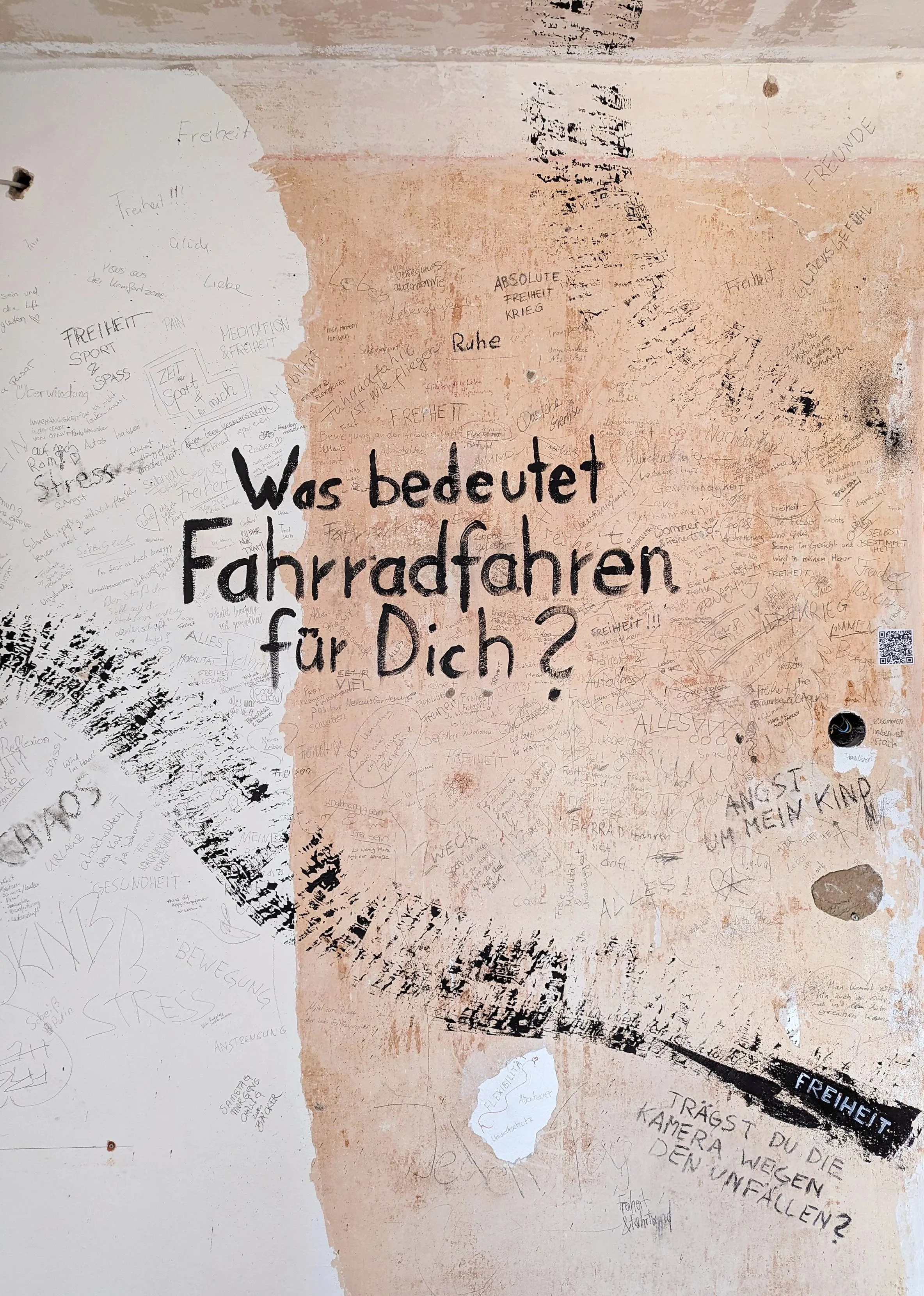 Wall with writing in German questions and comments, including a large text asking 'What does cycling mean to you?', surrounded by smaller handwritten notes and scribbles in different shades and styles.