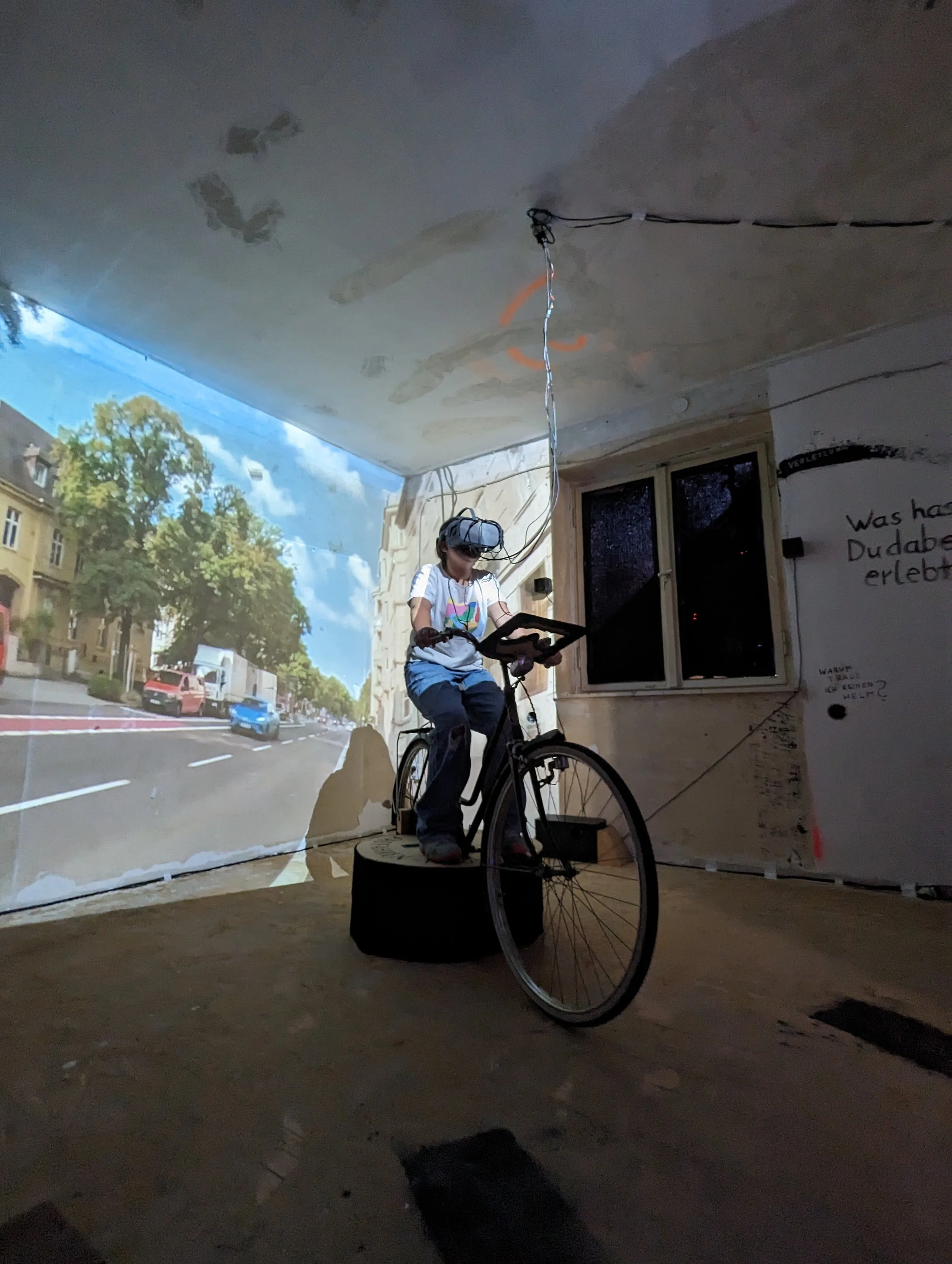A person wearing a virtual reality headset riding a stationary bicycle on a circular platform inside a room. The room has walls with projected images of a city street, and there is writing on the wall.