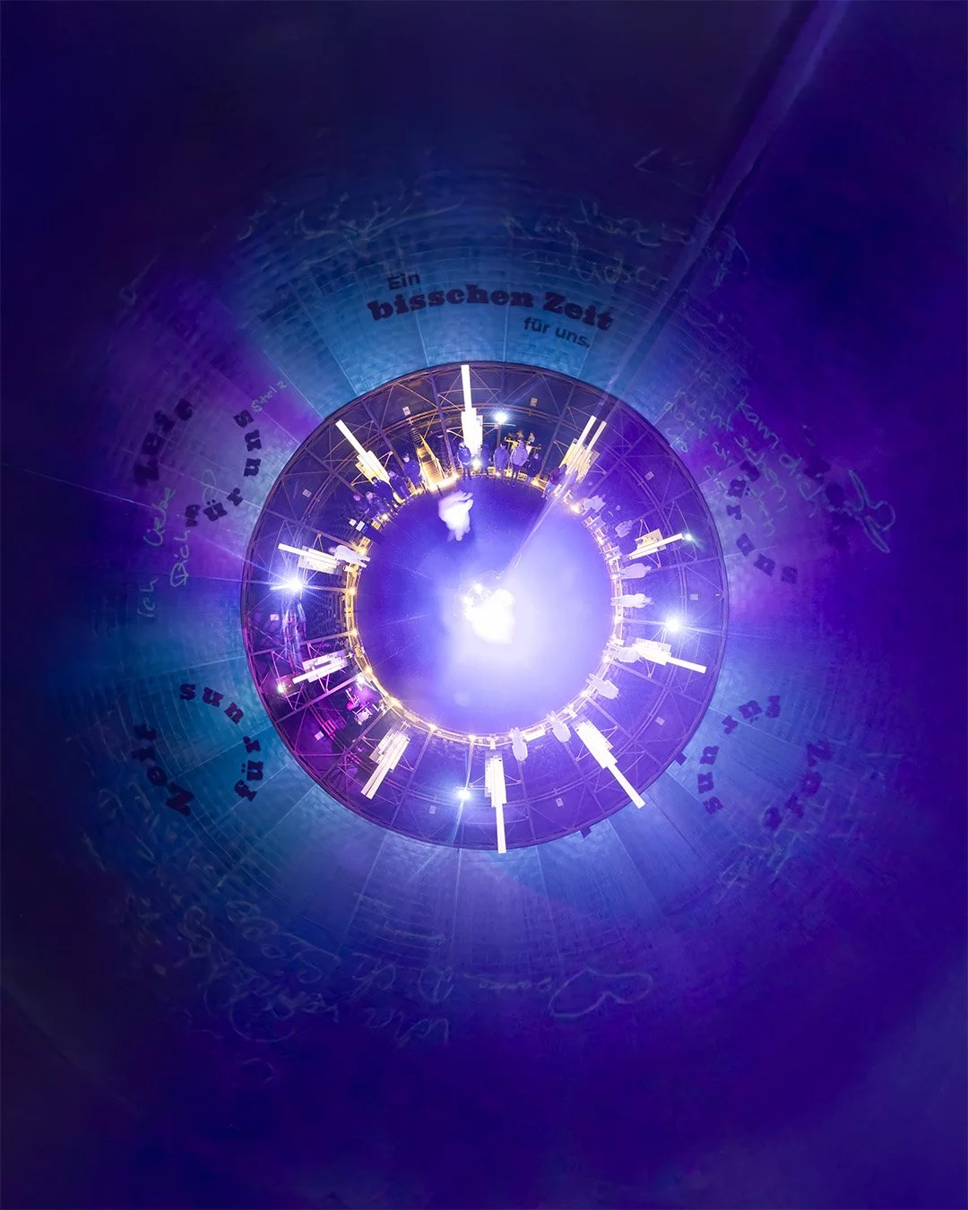 A brightly lit, circular clock-like installation with luminescent hands and numbers, viewed from below with colorful purple and blue lighting.