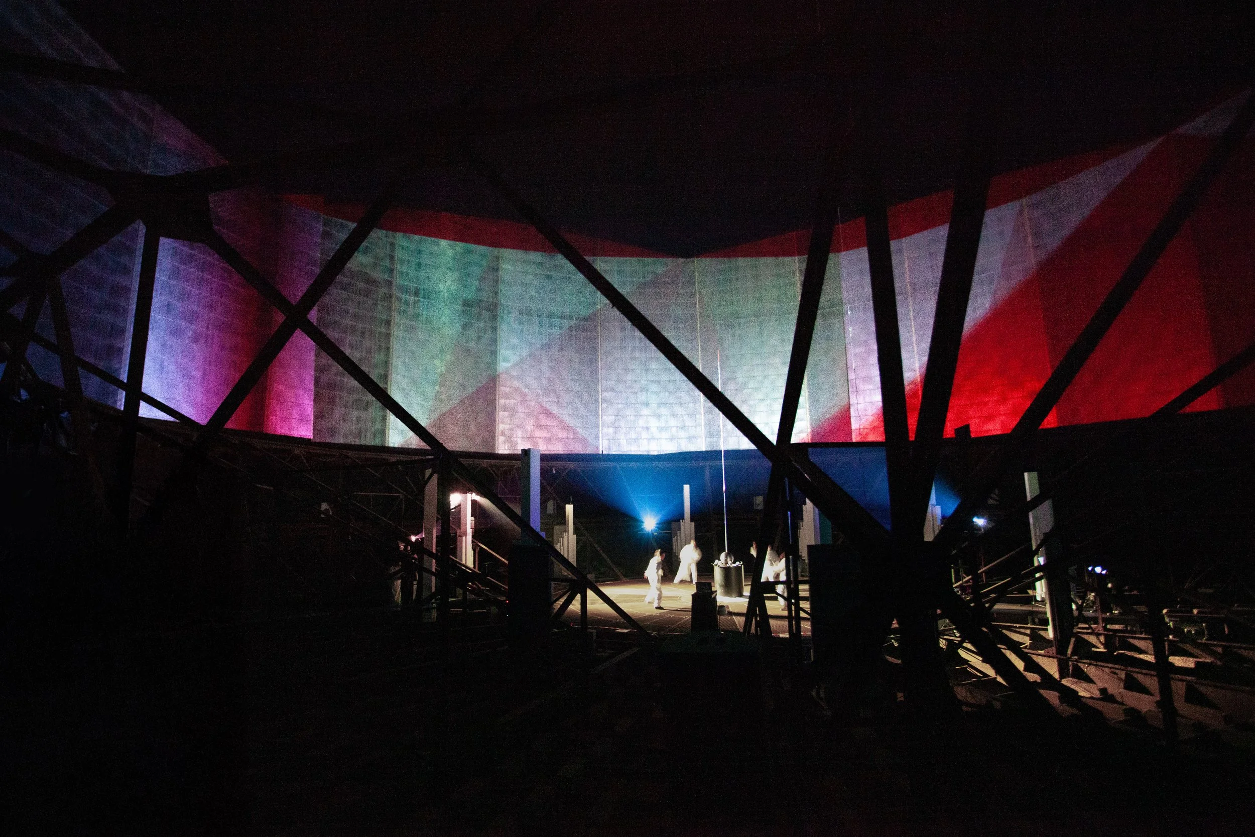 Wide view of performers inside a historic gasometer with large-scale projection mapping across the walls, creating an immersive theatre environment.