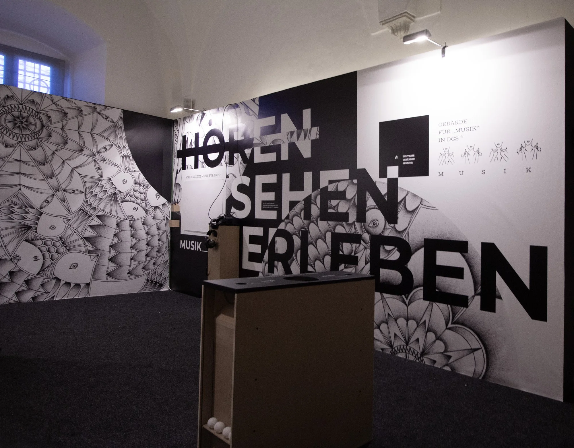 Exhibition room with black and white graphic wall art, large bold text, and a small wooden stand holding white balls.