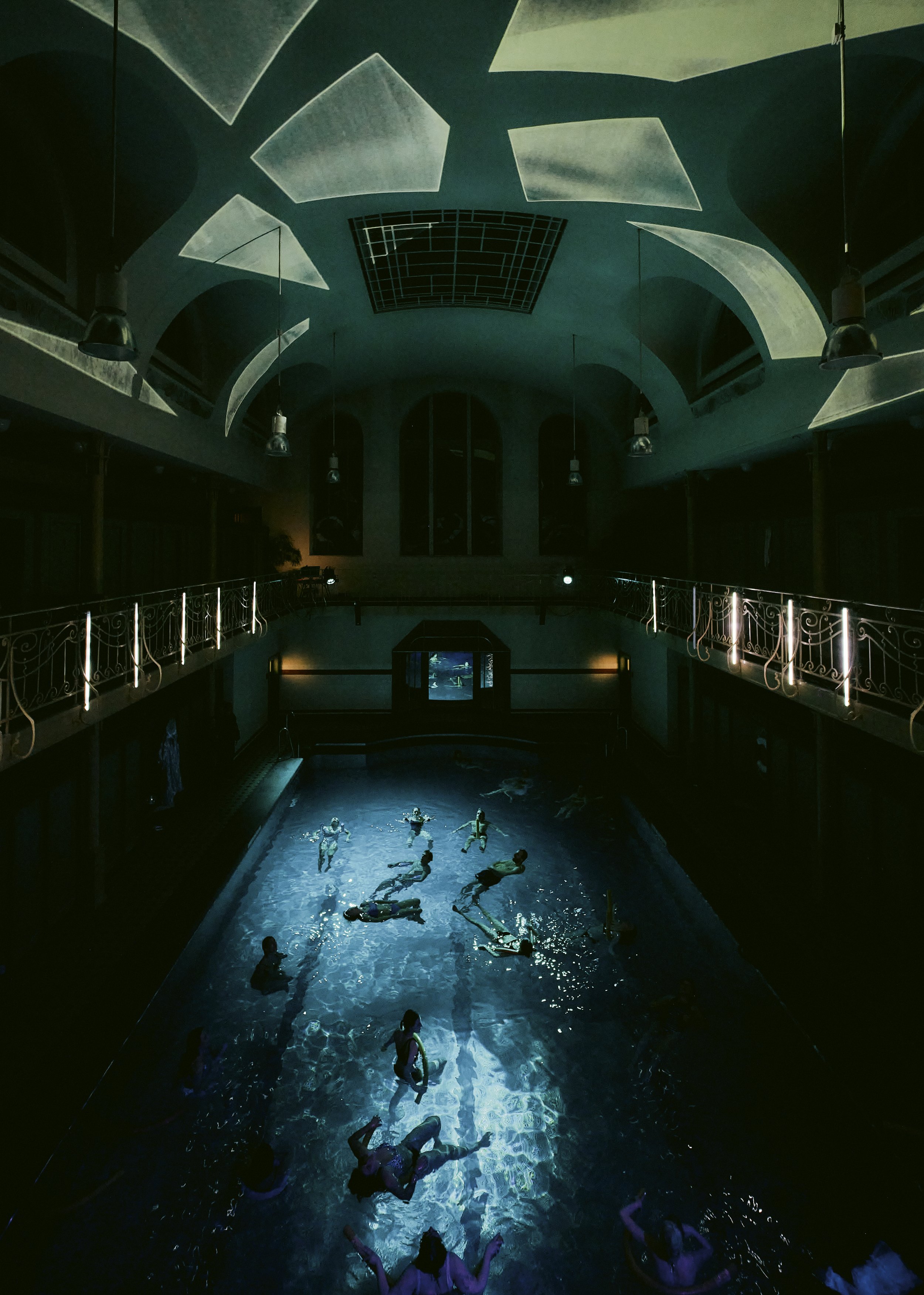People swimming and floating in a historic bathhouse, with the ceiling partially illuminated and a balcony with ornate railings.