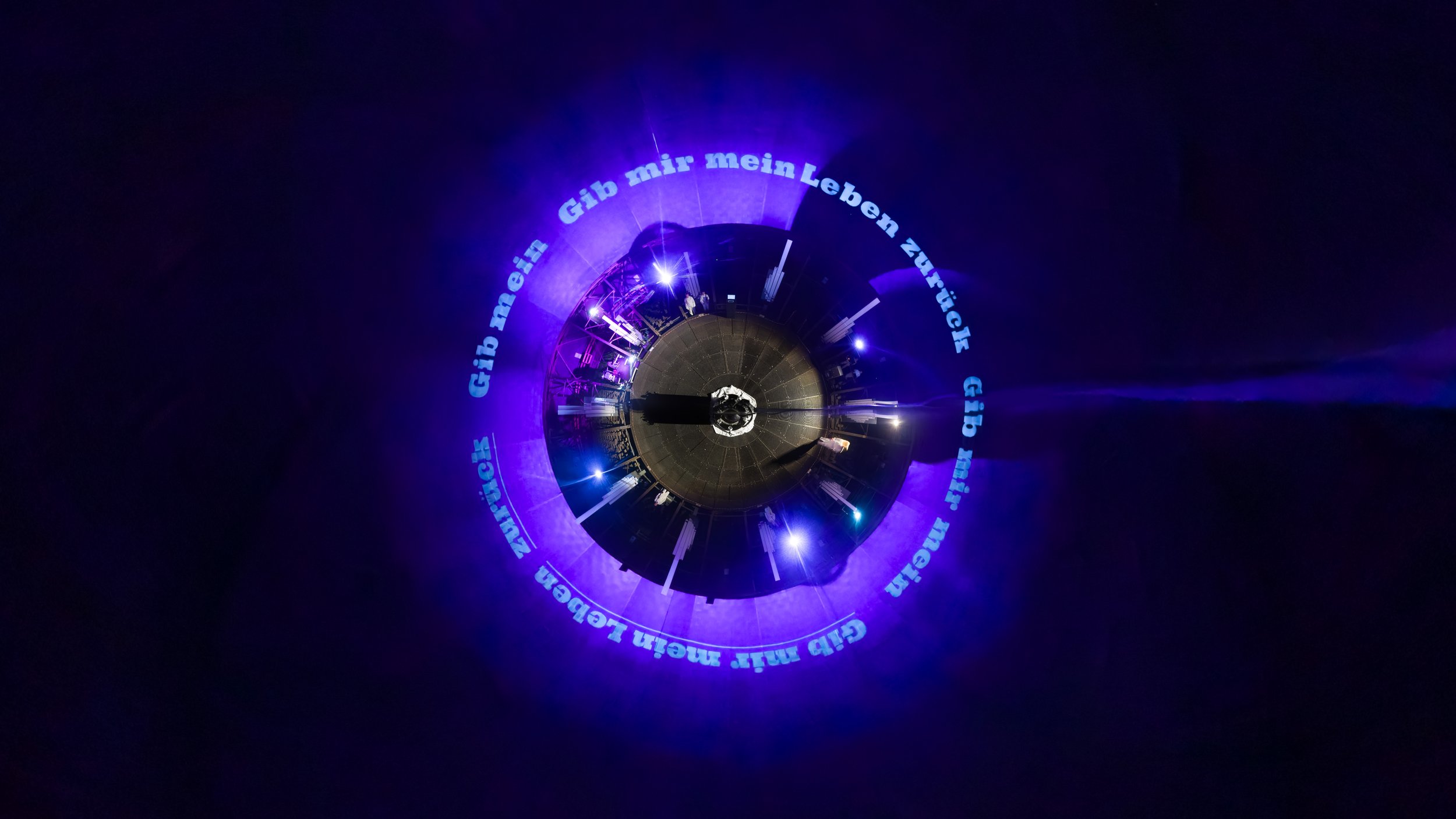 A circular stage with purple lighting and a ring of German text that says "Give me my life back" wrapping around the gasometer walls, synchronized with live music during an immersive theatre performance.