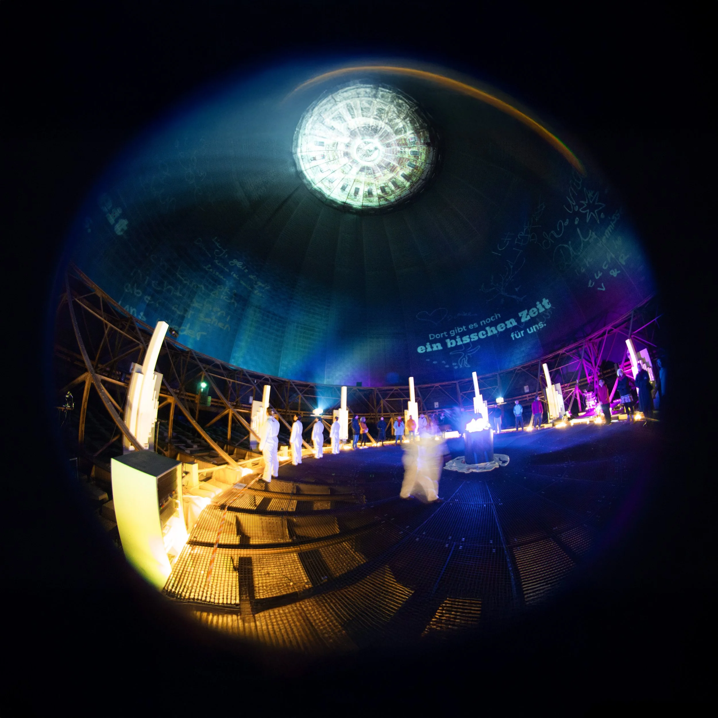 Wide-angle view inside a historic gasometer with immersive 360° projections, performers and audience surrounded by light and architecture.
