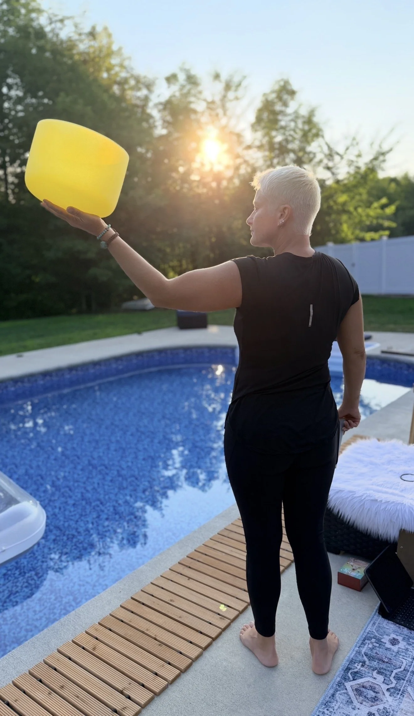 A woman with short blonde hair standing by a swimming pool during sunset, holding a large yellow bowl or vessel. She is dressed in black athletic clothing, barefoot, with a laptop and a box nearby on the deck.