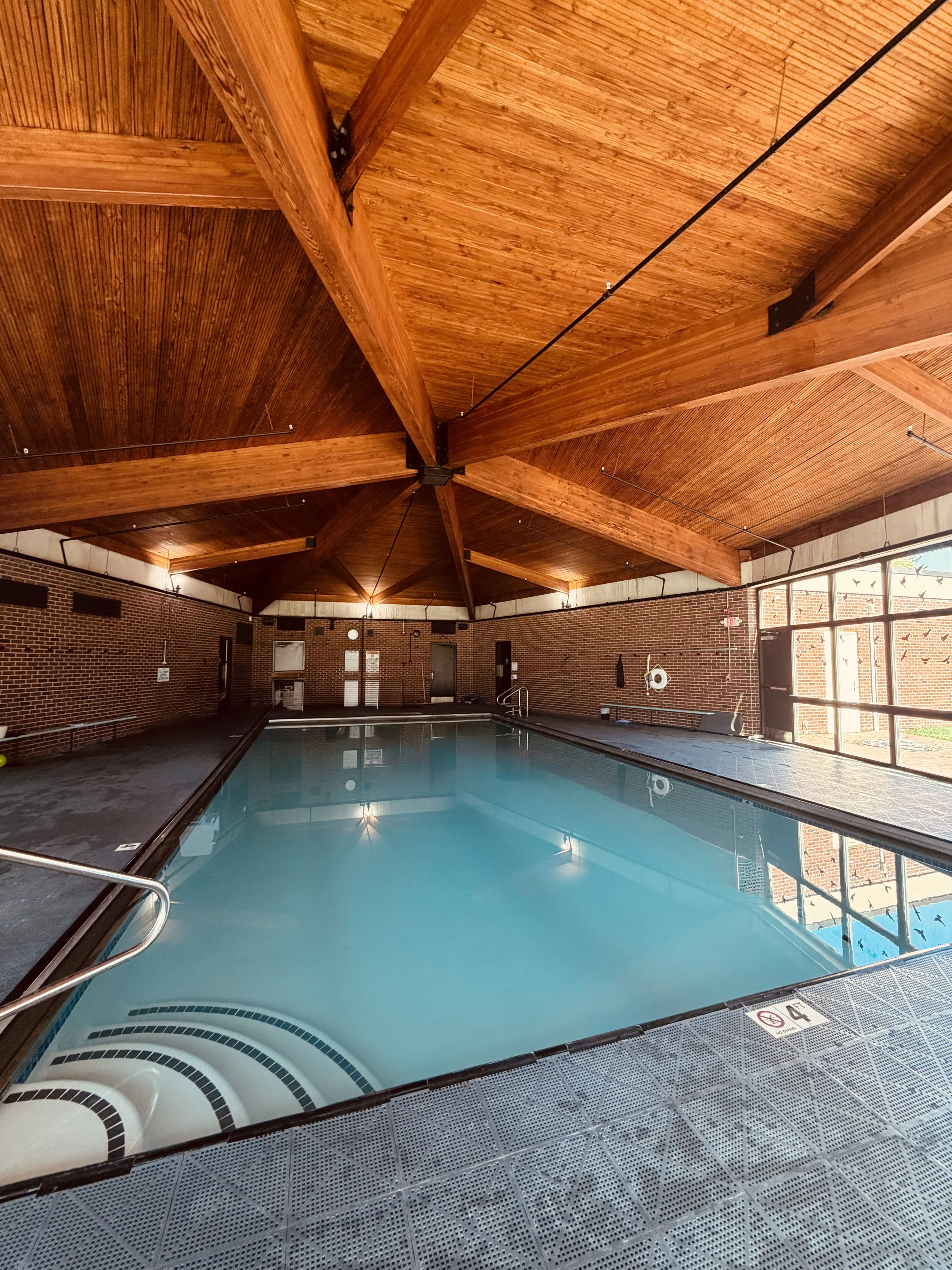 Indoor swimming pool with a wooden ceiling and brick walls, large windows, and metal railings.
