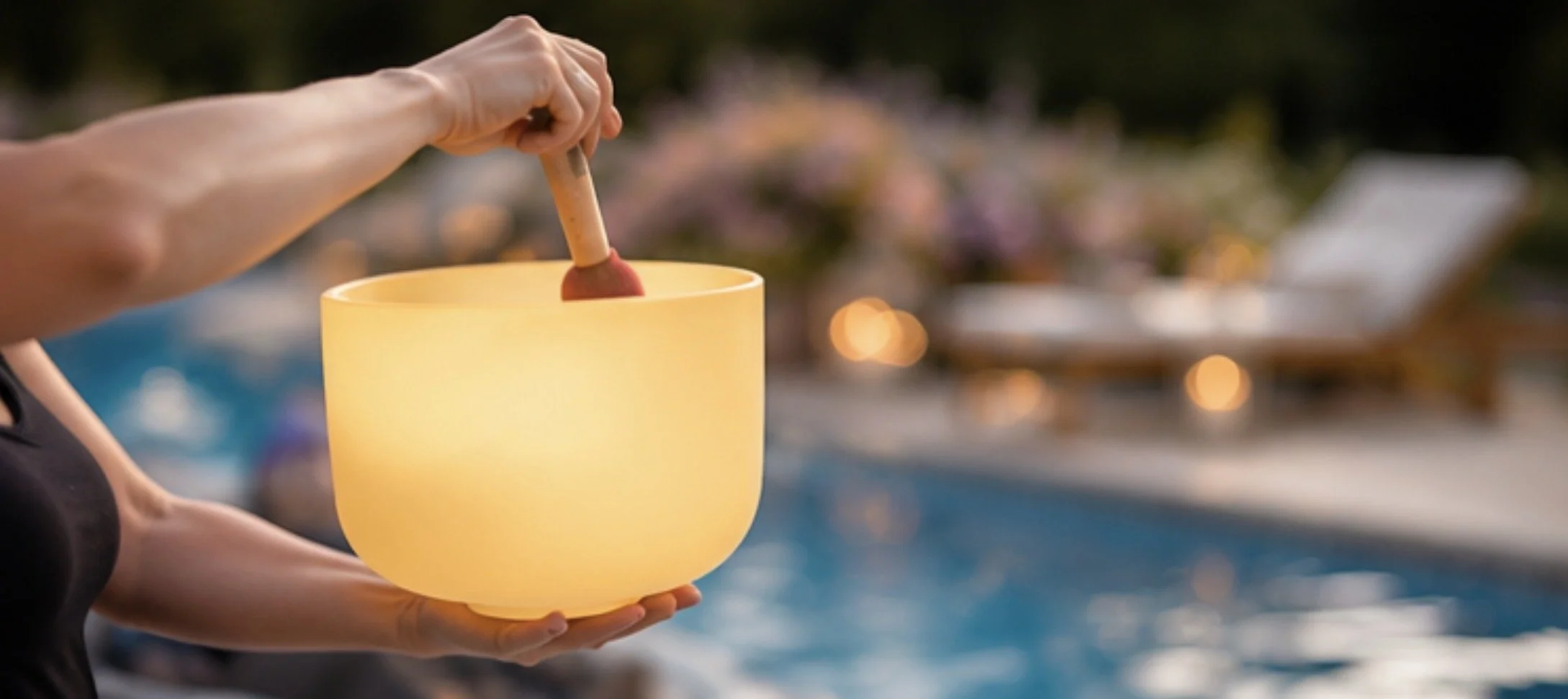 Person holding a yellow singing bowl with a mallet at a poolside setting during twilight.