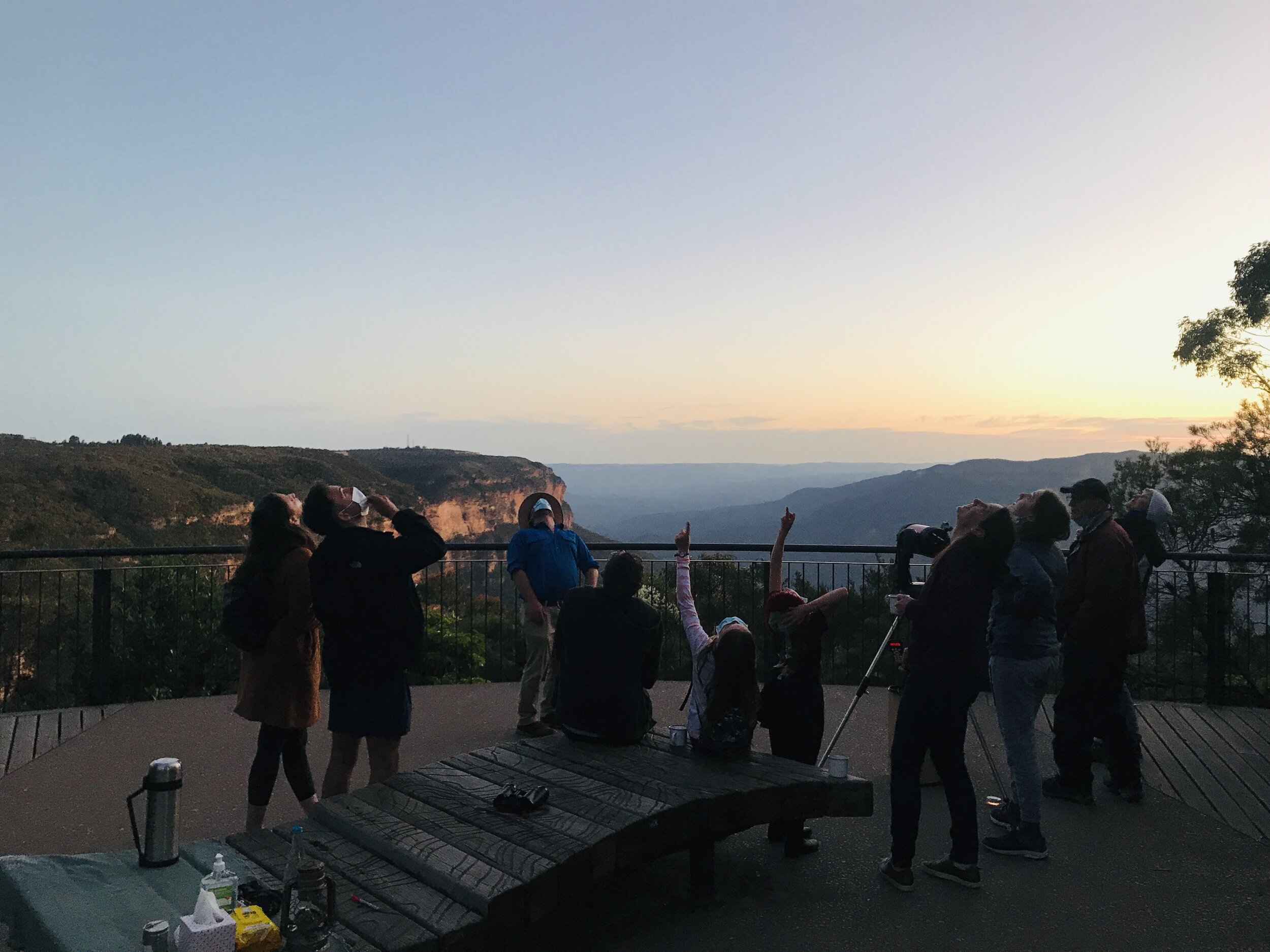 A group of people observing the sunset from a lookout point in a mountainous area, some using telescopes, with scenic mountain views in the background.