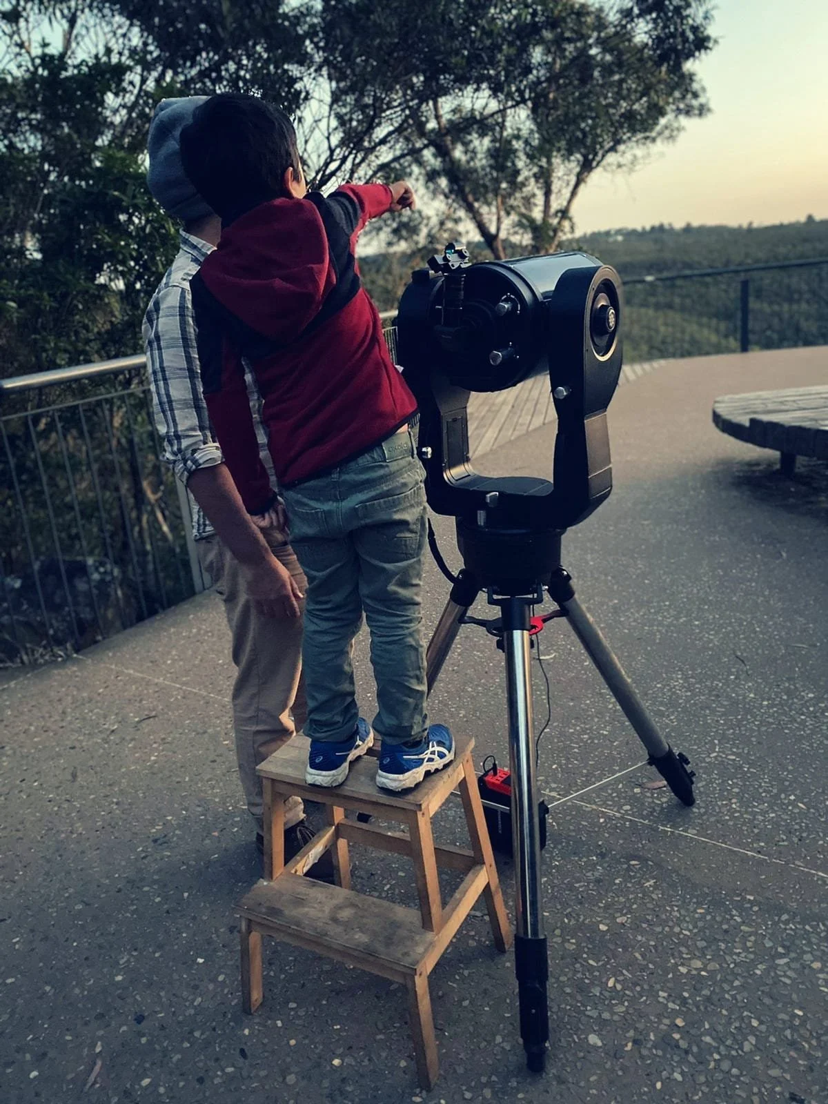 Two children observe the night sky with a telescope on a paved outdoor area at sunset, surrounded by trees and a railing.