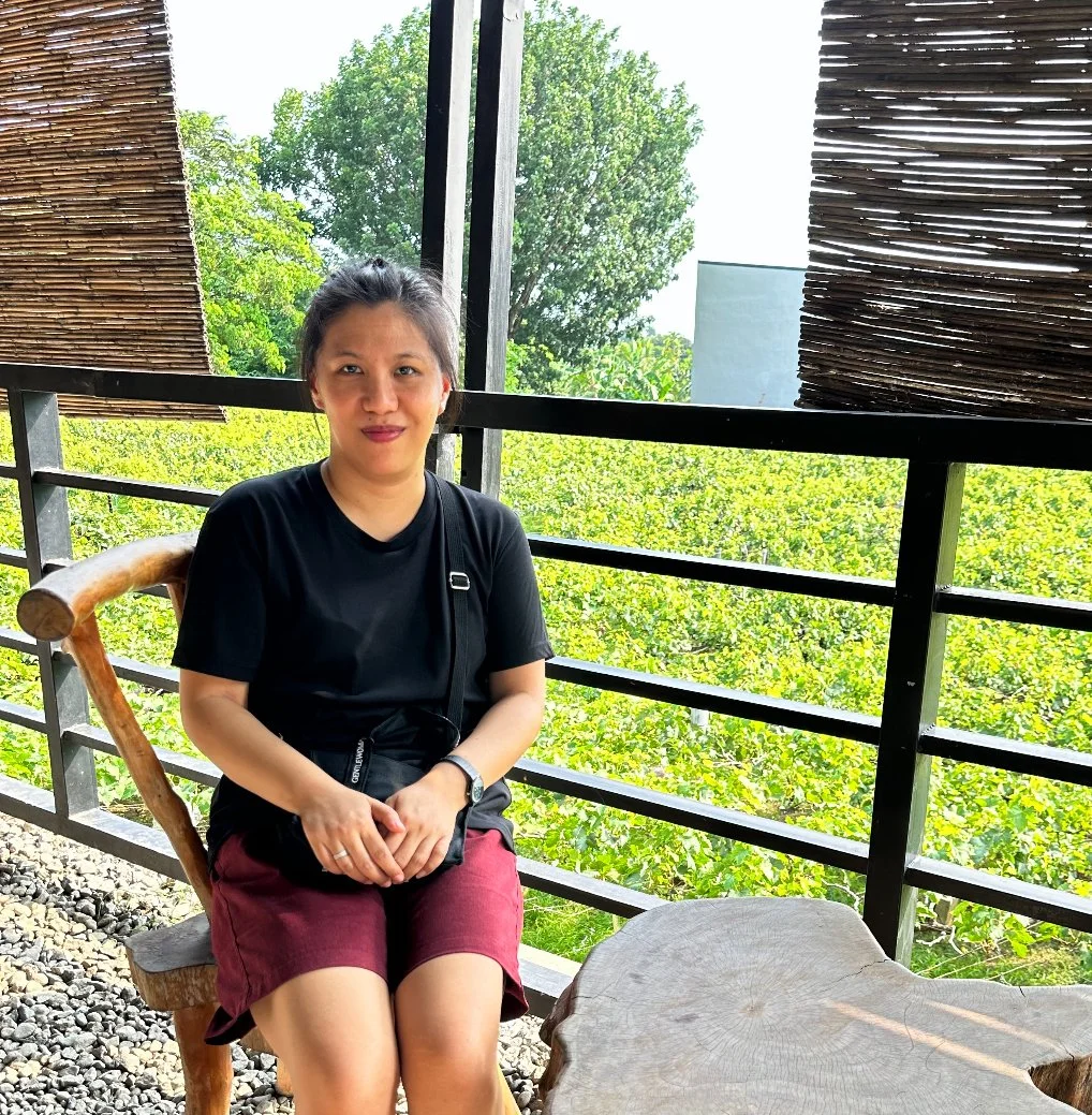 A woman sitting on a wooden chair on a balcony with trees in the background.