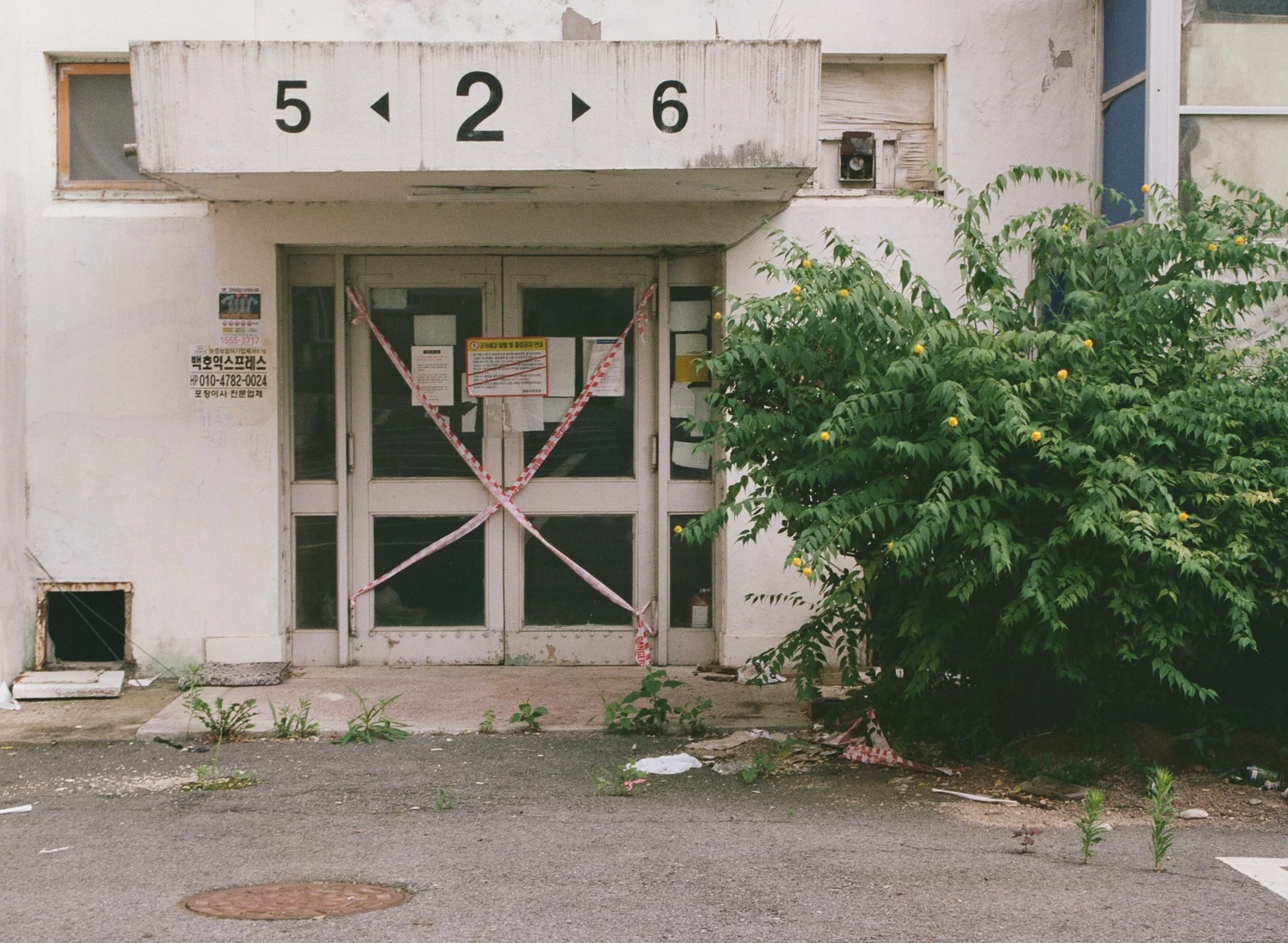 An abandoned building entrance with a door, a window, and a bush on the right. The building's facade is worn and has emergency tape across the door and window. Above, there is a sign with numbers 5, 2, and 6 and arrows pointing left and right.