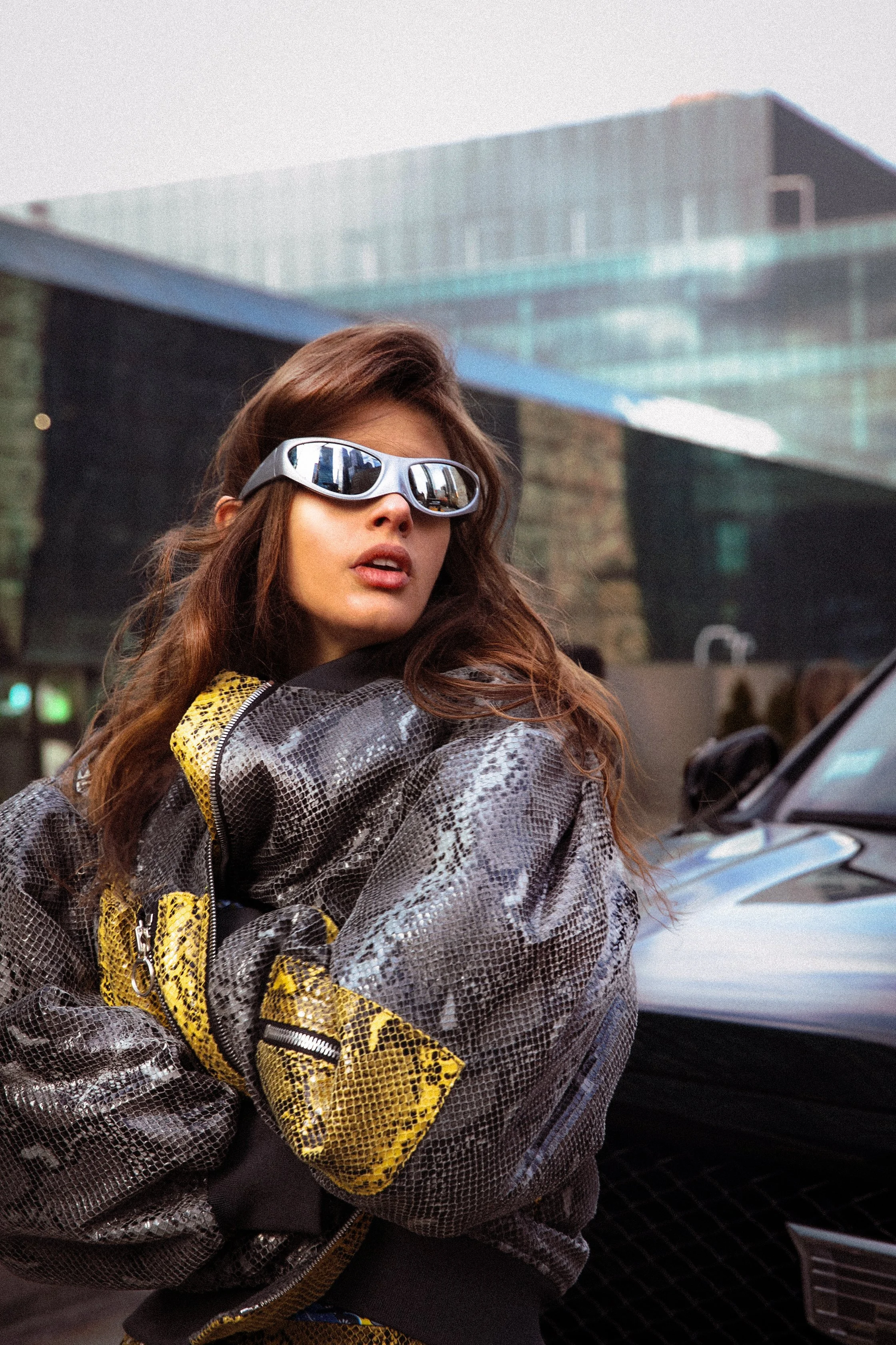 Young woman wearing reflective sunglasses and a snake print jacket, standing in front of a modern building and a vehicle.