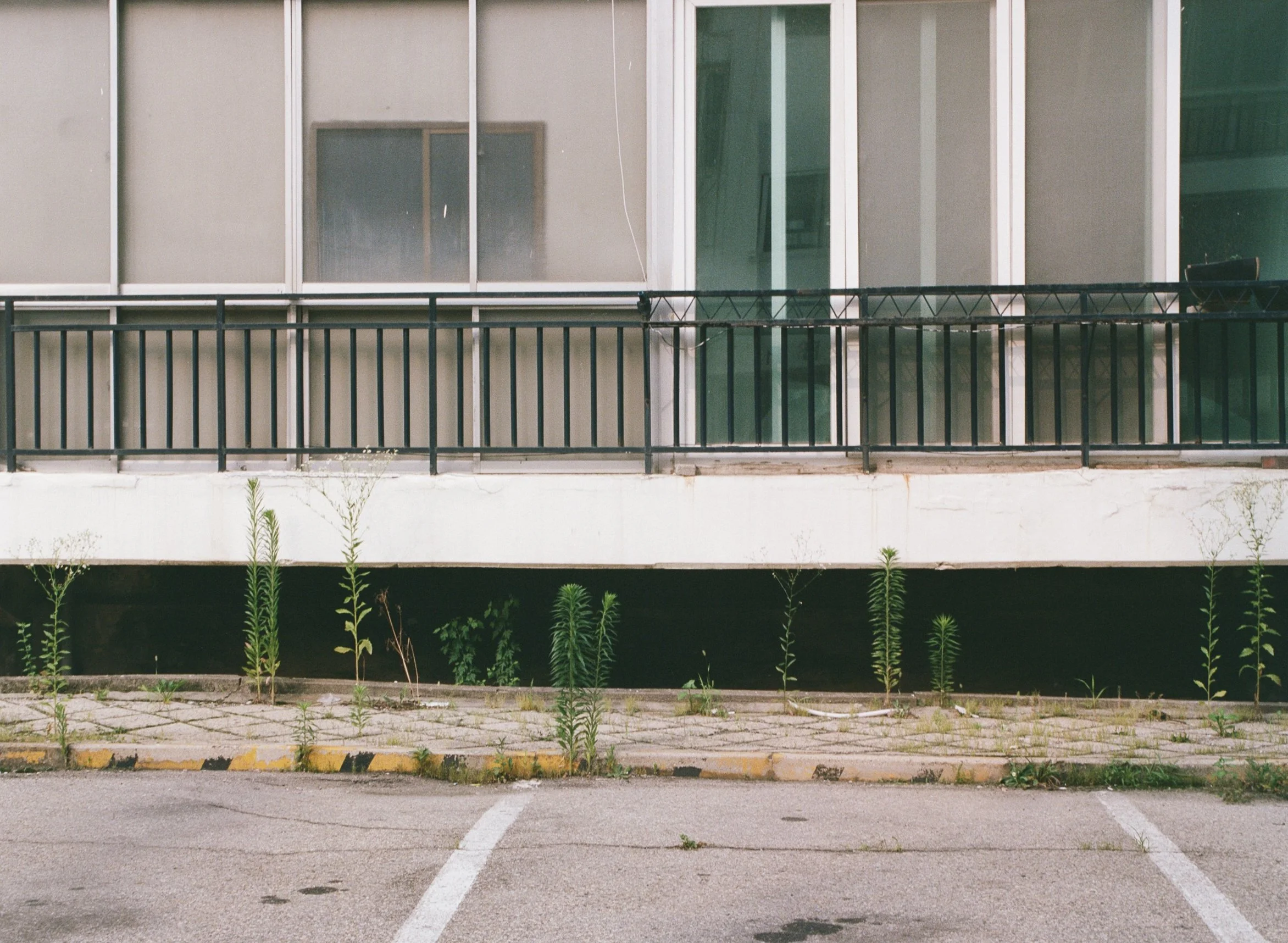 Empty parking lot with weeds growing between pavement cracks, in front of a modern building with large glass windows and a black metal railing on a balcony.