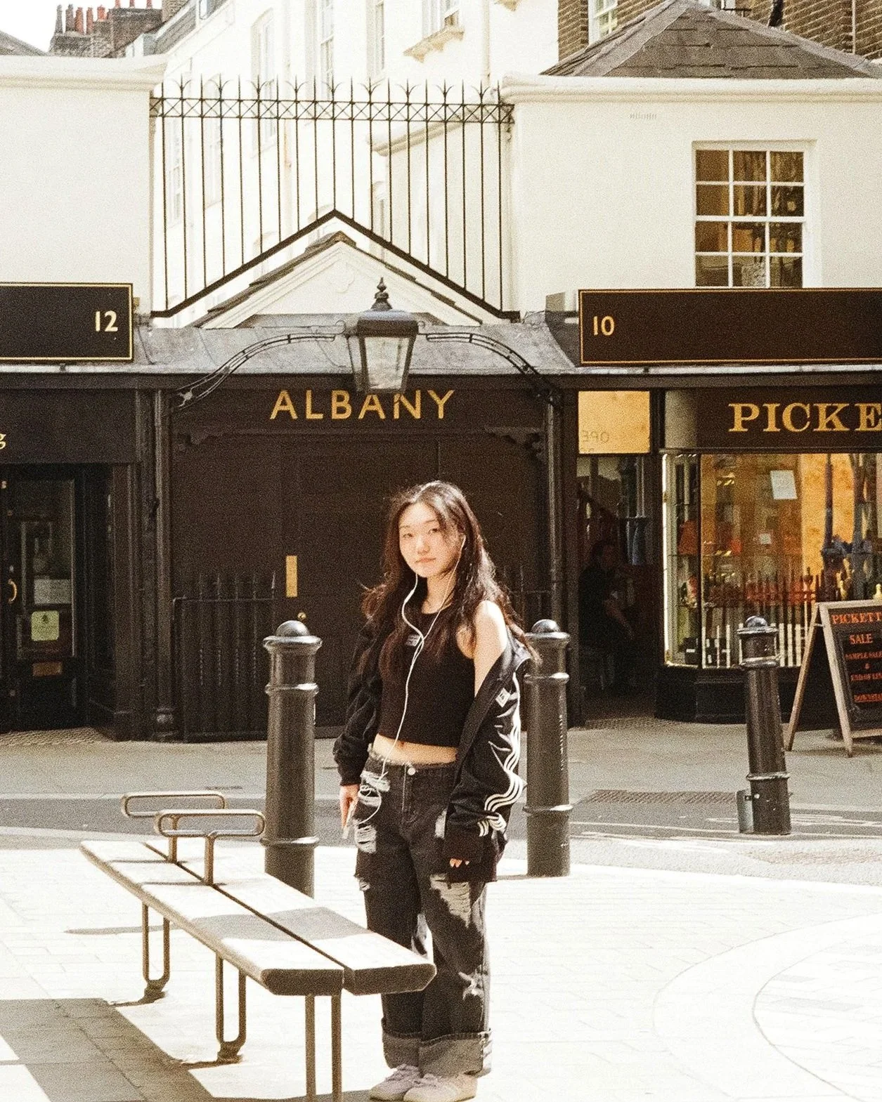 A young woman with long dark hair wearing a black crop top, black jacket, and ripped jeans standing on a city sidewalk in front of a black storefront with the word 'Albany' on it, with a bench and black bollards nearby.