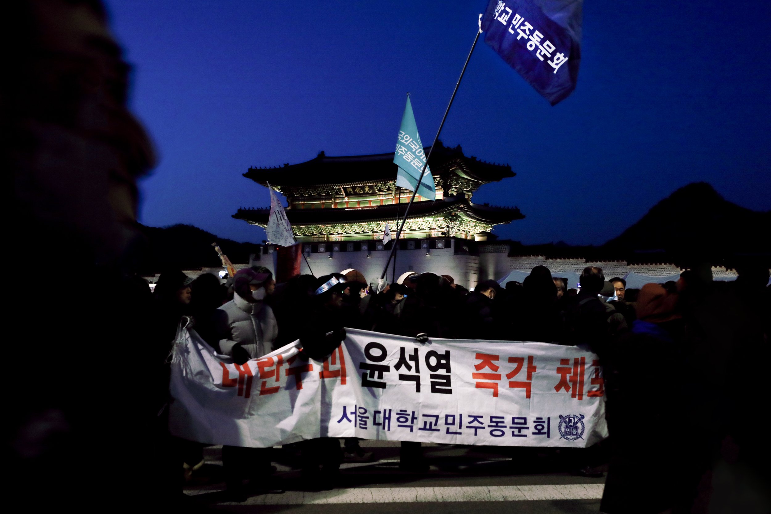 People gathered at a night protest in front of Gyeongbokgung Palace, holding banners and flags. Some individuals are wearing face masks, and the flags display Korean text. The crowd is illuminated by artificial lighting, with the palace’s traditional architecture lit up in the background.
