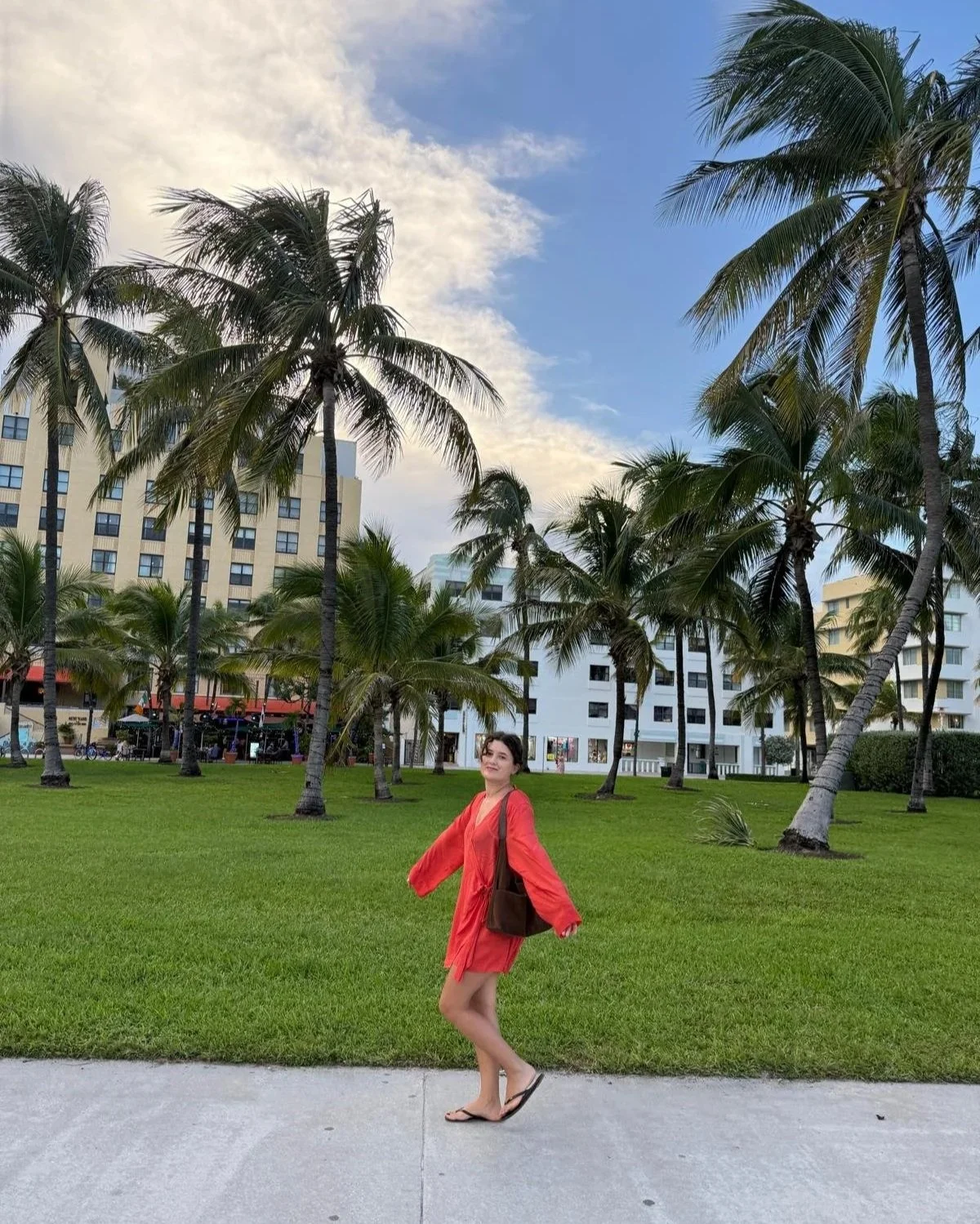A woman wearing a red dress and sandals standing on a sidewalk in front of a grassy area with palm trees and buildings in the background under a partly cloudy sky.