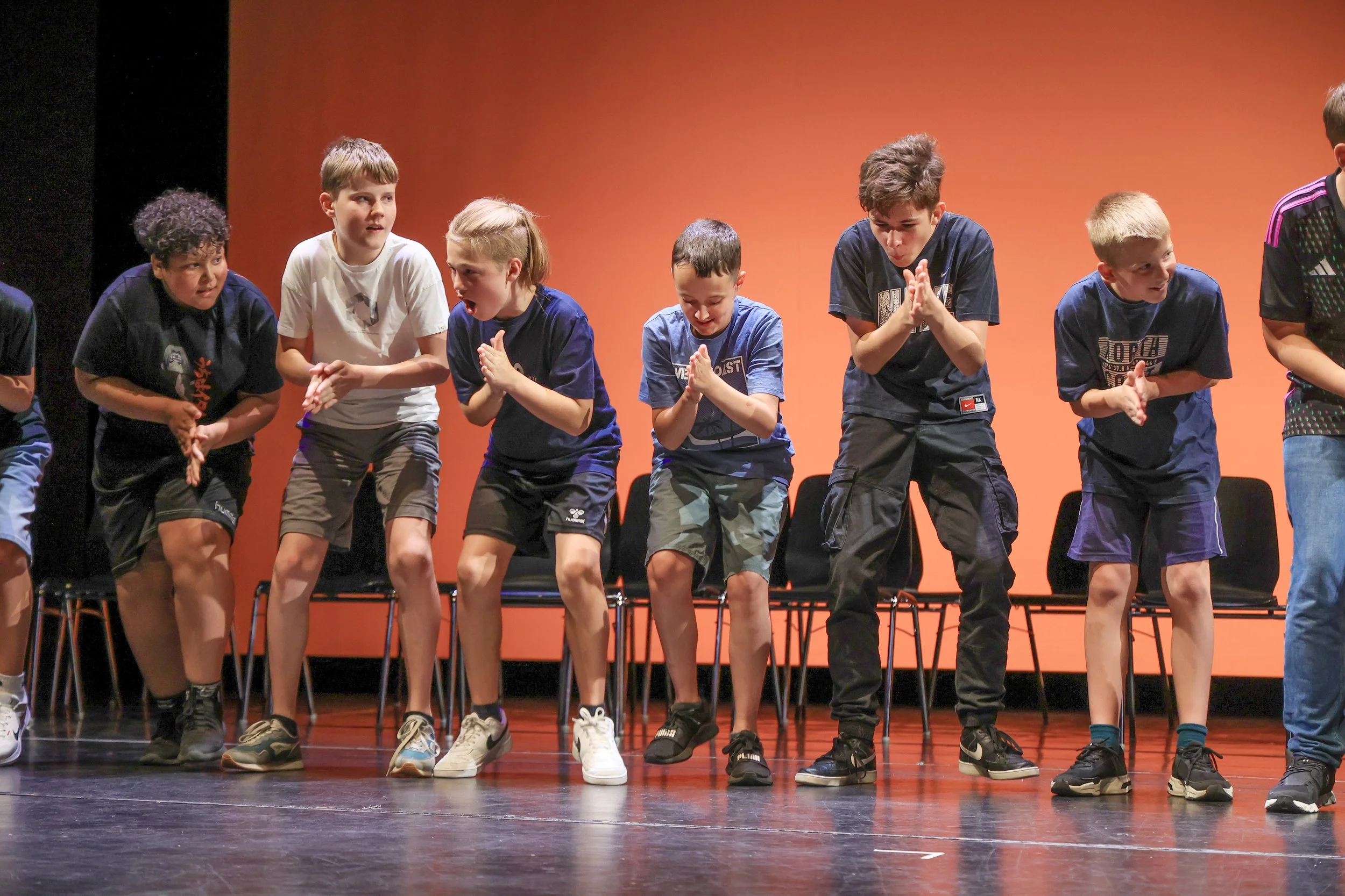 Junge Kinder beim Tanz auf einer Bühne, verstellt mit Stühlen, orange Hintergrund.