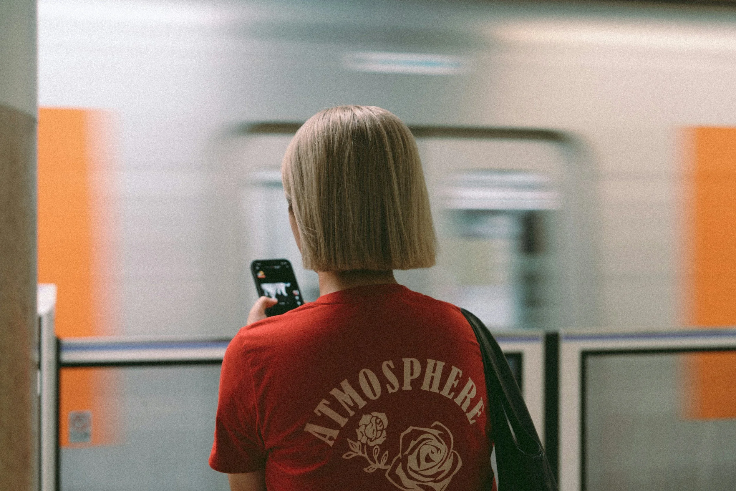 A woman with short blonde hair wearing a red t-shirt with the word 'ATMOSPHERE' and floral design on the back, standing in an indoor setting and looking at her phone.