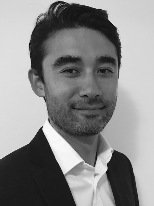 Black and white photo of a man with dark hair, light beard, wearing a suit jacket and white collared shirt, smiling slightly, standing against a plain background.