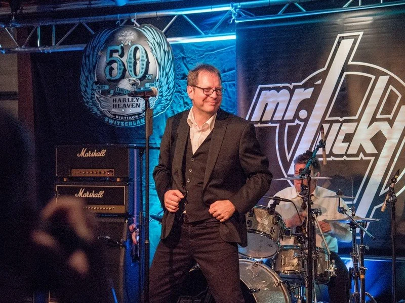 Ein Mann trägt Anzug auf einer Bühne mit Schlagzeug und Verstärkern, im Hintergrund ein Banner mit 'mr. lucky' und ein weiteres Banner mit '50' Jahrestag der Harley-Davidson in der Schweiz.