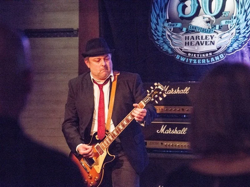 Ein Mann spielt E-Gitarre bei einer Veranstaltung, im Hintergrund ein Banner mit dem Harley-Heaven-Logo und einigen Lautsprechern.