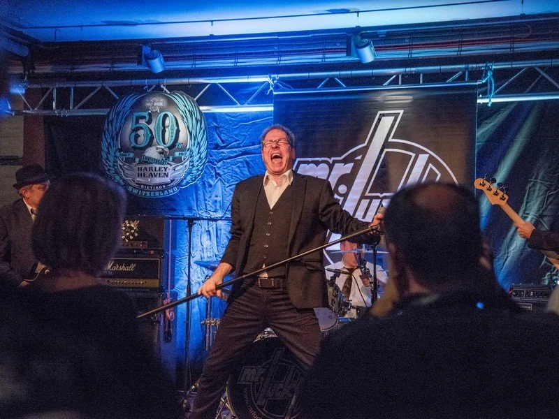 Ein Mann singt und spielt mit einem Mikrofonstab bei einer Live-Musikveranstaltung, umgeben von Musikern und einem Publikum, mit einem Banner im Hintergrund, das den 50. Jahrestag eines Harley-Davidson Jubiläums zeigt.