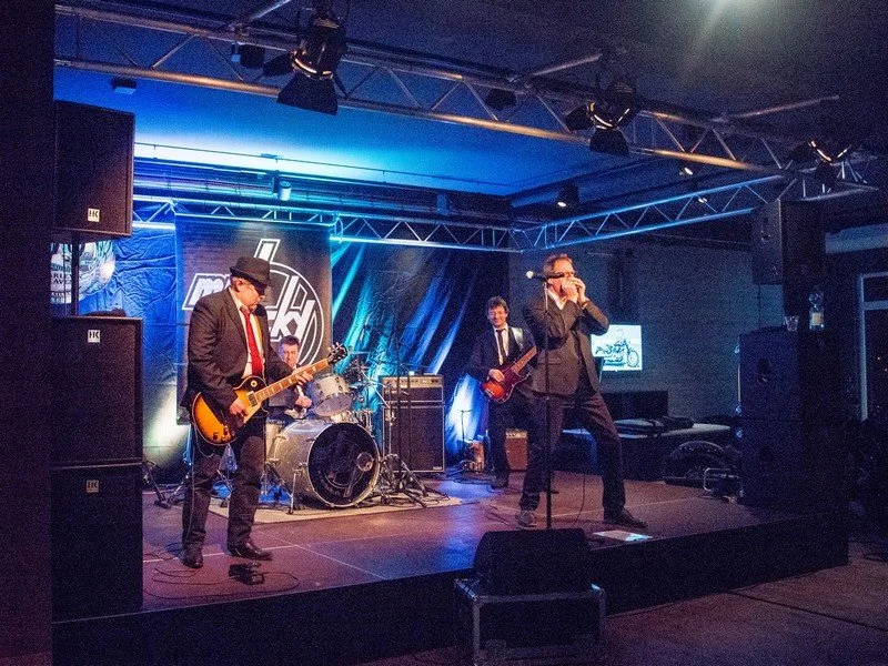 Eine Band spielt auf einer Bühne bei einer Live-Performance, bestehend aus einem Gitarristen, einem Bassisten, einem Schlagzeuger und einem Sänger, in einem dunklen, professionellen Veranstaltungsraum.