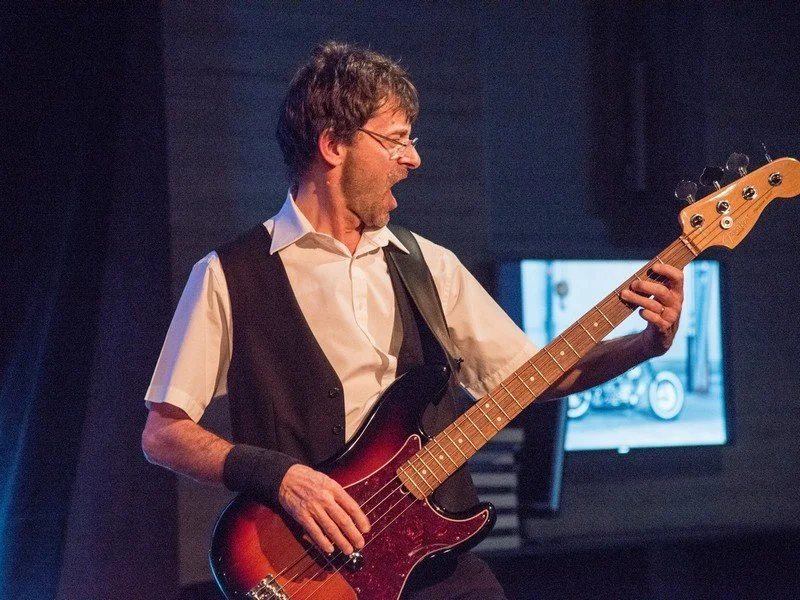 Ein Mann spielt Bassgitarre auf einer Bühne, trägt eine weiße Hemd, schwarzen Weste und Armband, und hat kurze braune Haare und Brille.