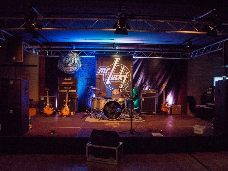 Bühne mit Musikinstrumenten, darunter ein Schlagzeug, drei E-Gitarren, und Verstärker, mit einem magischen Hintergrund zentrum, das das Logo "mr. duck" zeigt, und einer Gedenkballon mit der Zahl 50.