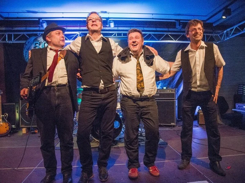 Vier Männer mit Mikrofonen und Musikinstrumenten feiern auf einer Bühne, umgeben von Musiktechnik.