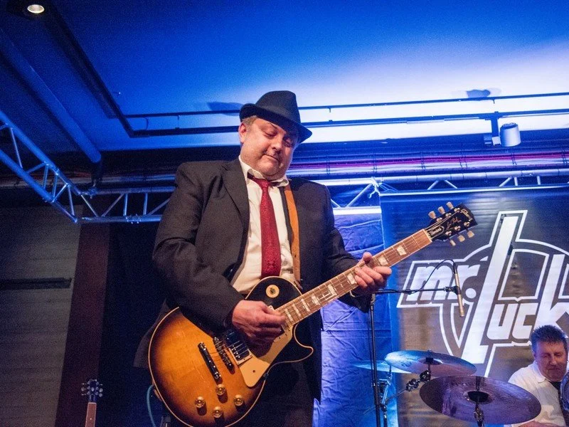 Ein Mann im schwarzen Anzug, Hemd und Hut spielt Gitarre auf einer Bühne bei einem Konzert.