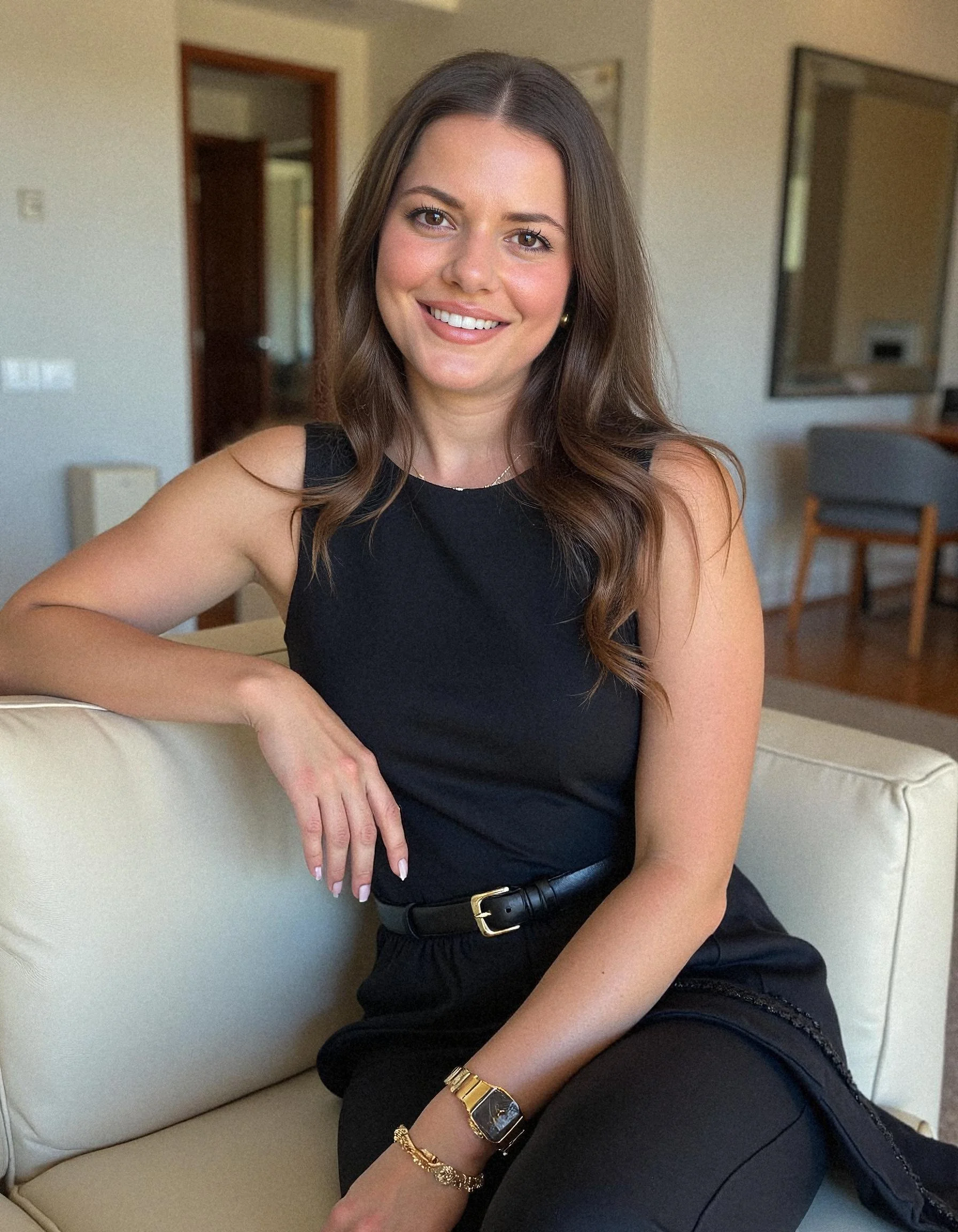 A young woman with long wavy brown hair smiling while sitting on a cream-colored couch in a room with a door, mirror, and chairs in the background. virtual assistant RADOVANA