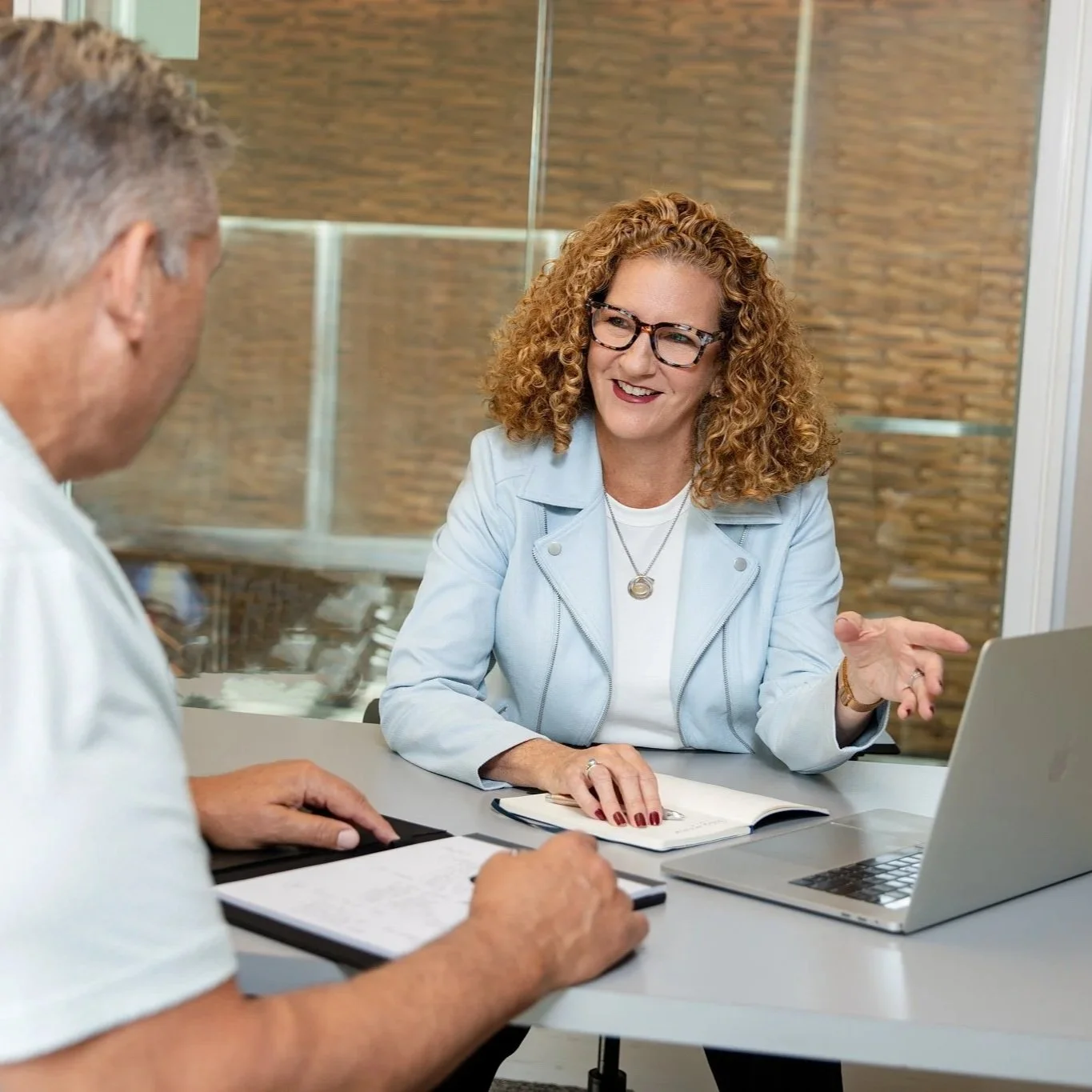 A woman with curly red hair, glasses, and a light blue jacket is smiling and talking to a man in a white shirt. They are sitting at a table with a laptop, notebook, and tablet, in a modern office setting with a glass wall and brick wall in the background.