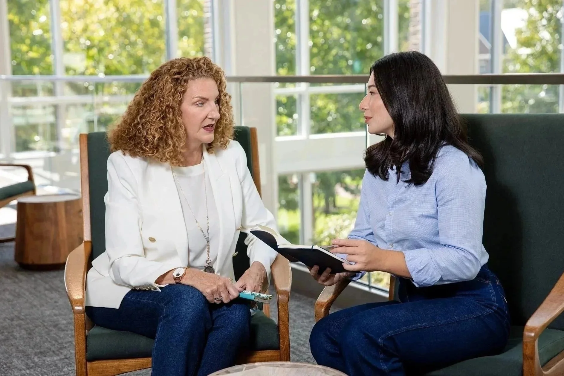 Two women sitting and talking in a room with large windows, one holding a notebook.