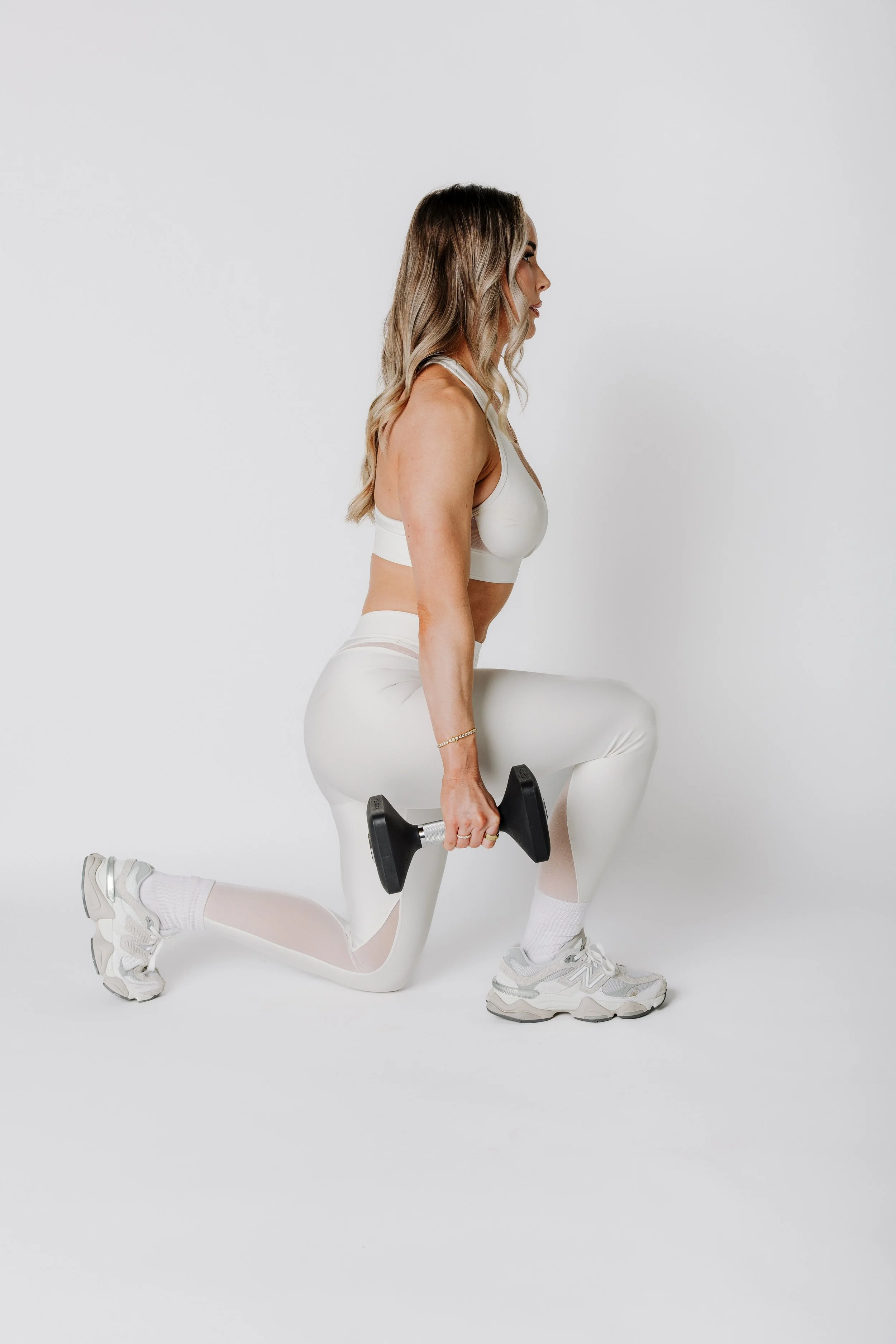A woman in white activewear performing a kneeling lunge exercise while holding a dumbbell in her right hand against a plain white background.