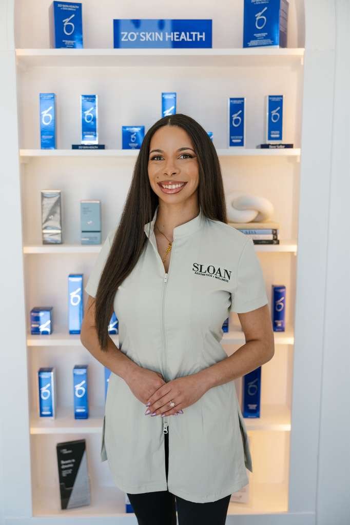 A smiling woman with long dark hair standing in front of a display shelf with skincare products. She is wearing a light-colored uniform with 'SLOAN' embroidered on it, and the background features a sign that reads 'ZO'SKIN HEALTH'. The products on the shelves are predominantly blue and white.