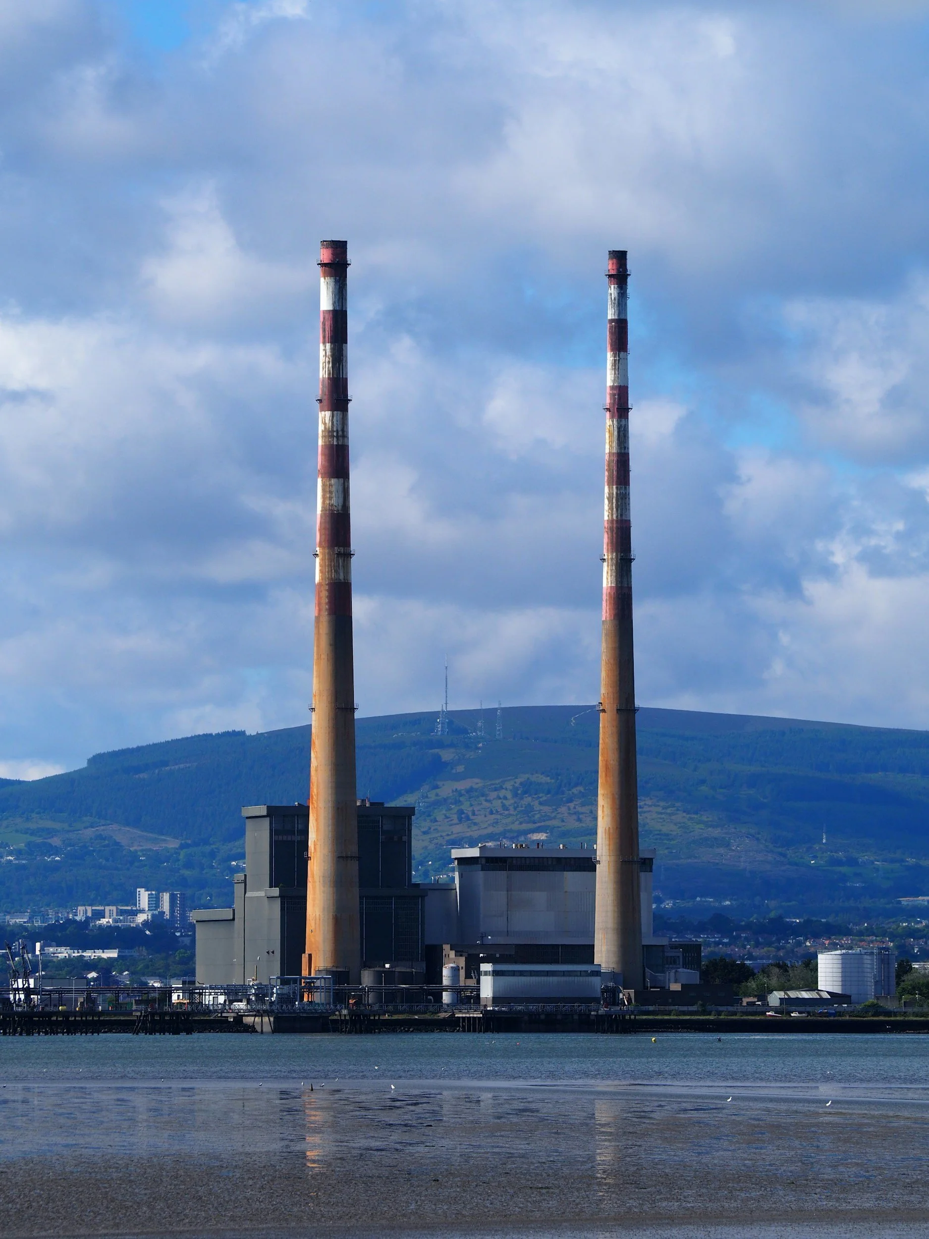 A power plant with two tall, striped smokestacks near water, with hills and a cloudy sky in the background.