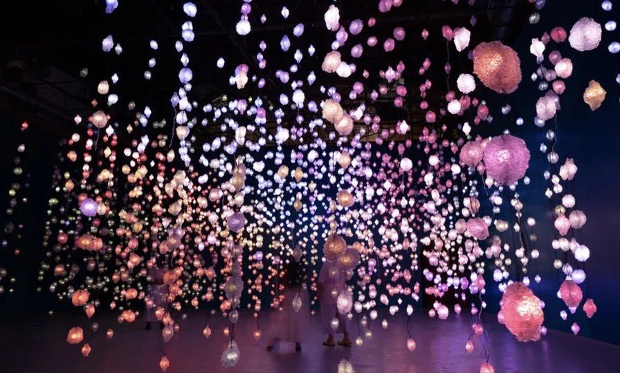 An art installation featuring numerous hanging globes and spheres illuminated with pink, purple, and white lights, creating a mesmerizing and colorful display on a dark background.