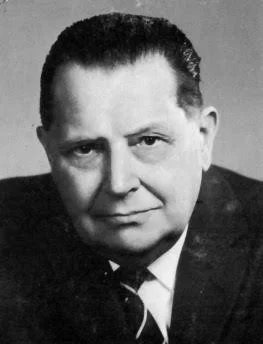 Black and white portrait of a serious-looking man with slicked-back hair, wearing a dark suit and tie.