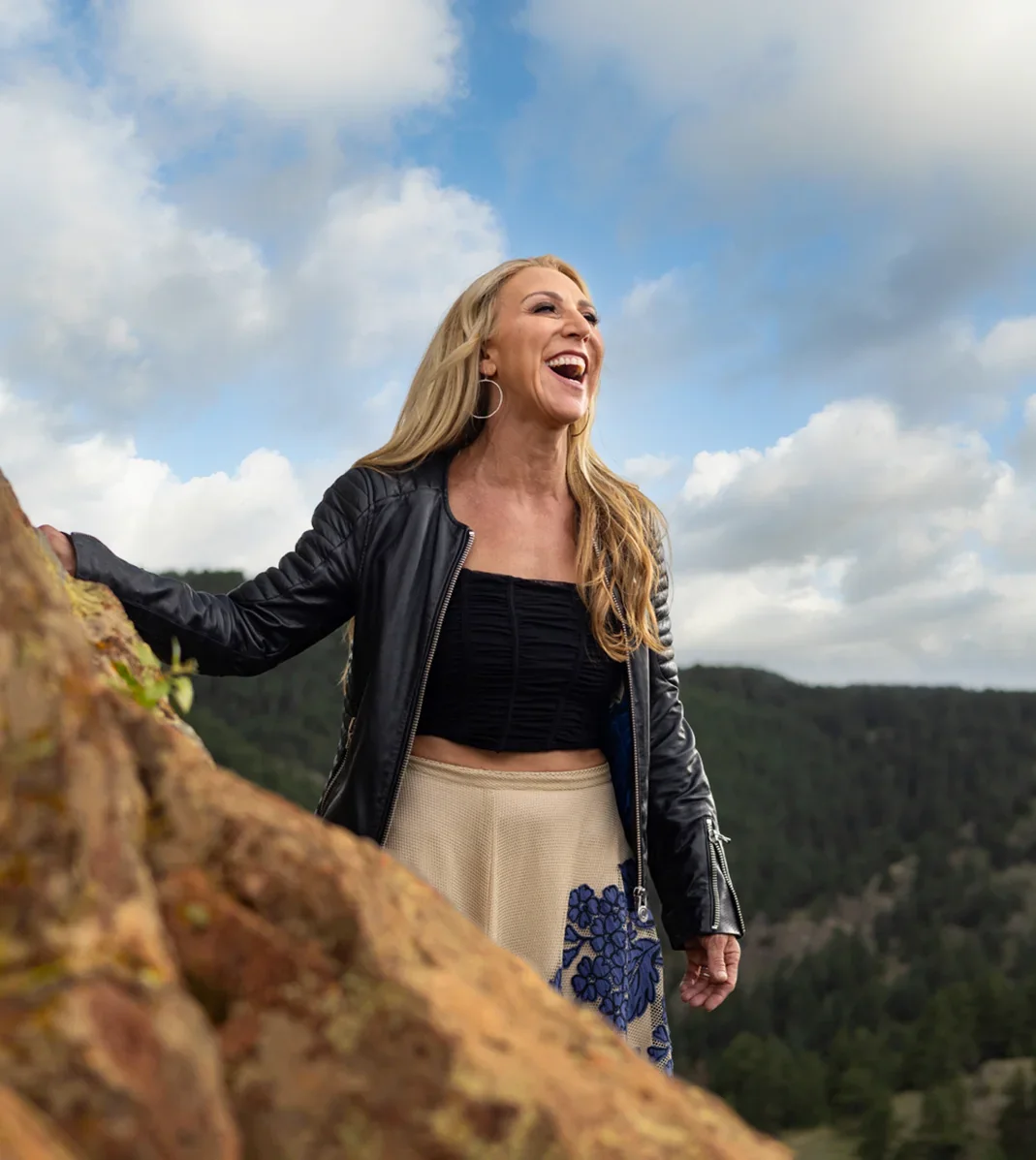 Lisa Fierer laughing outdoors on a mountain overlook, wearing a black jacket with a scenic forest backdrop.