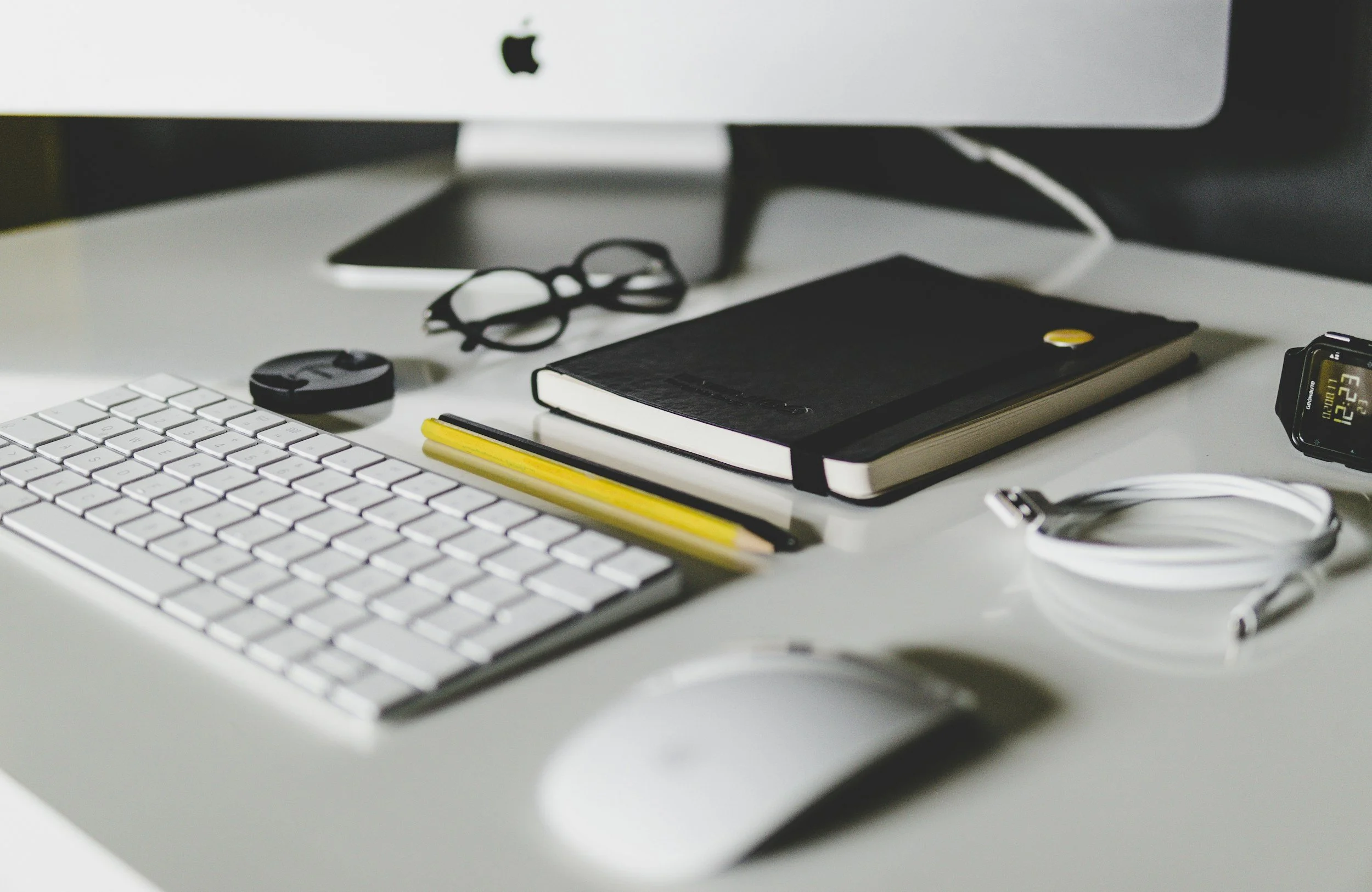 Schreibtisch mit Computer, Tastatur, Maus, Notizbuch, Brille, Kugelschreiber, Kabel und Smartwatch.
