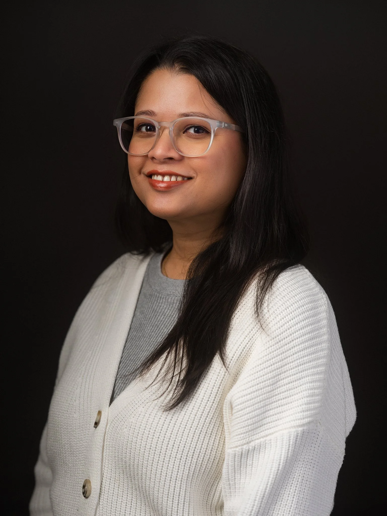 A young woman with dark hair, glasses, and small earrings, wearing a white shirt, standing in front of beige curtains.